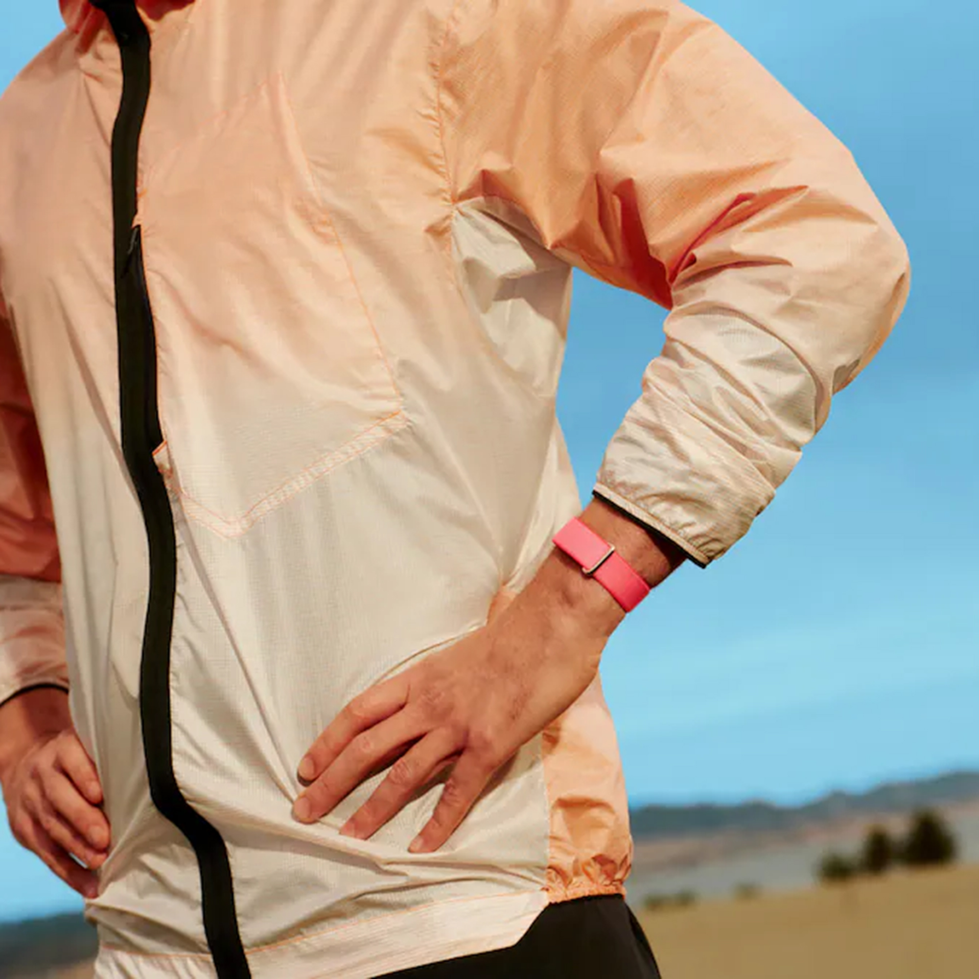 Persona con chaqueta rosa y pulsera rosa brillante, con cielo azul y paisaje de fondo.