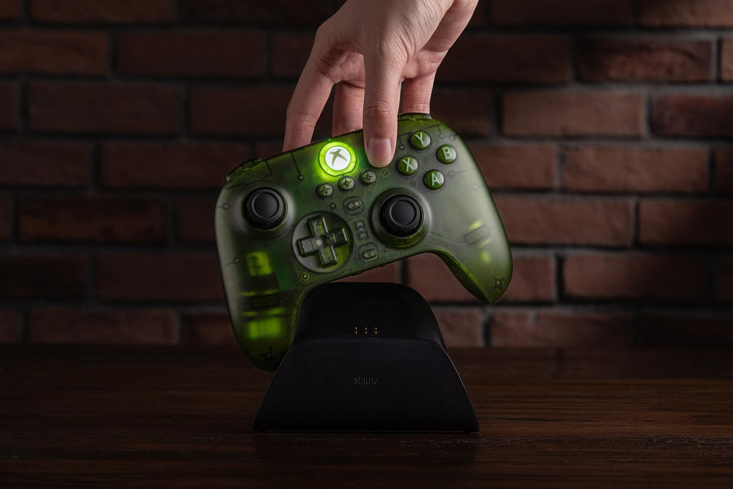 Ręka kładzie świecący, zielony, przezroczysty kontroler Xbox na czarnej stacji ładującej przed ceglaną ścianą.