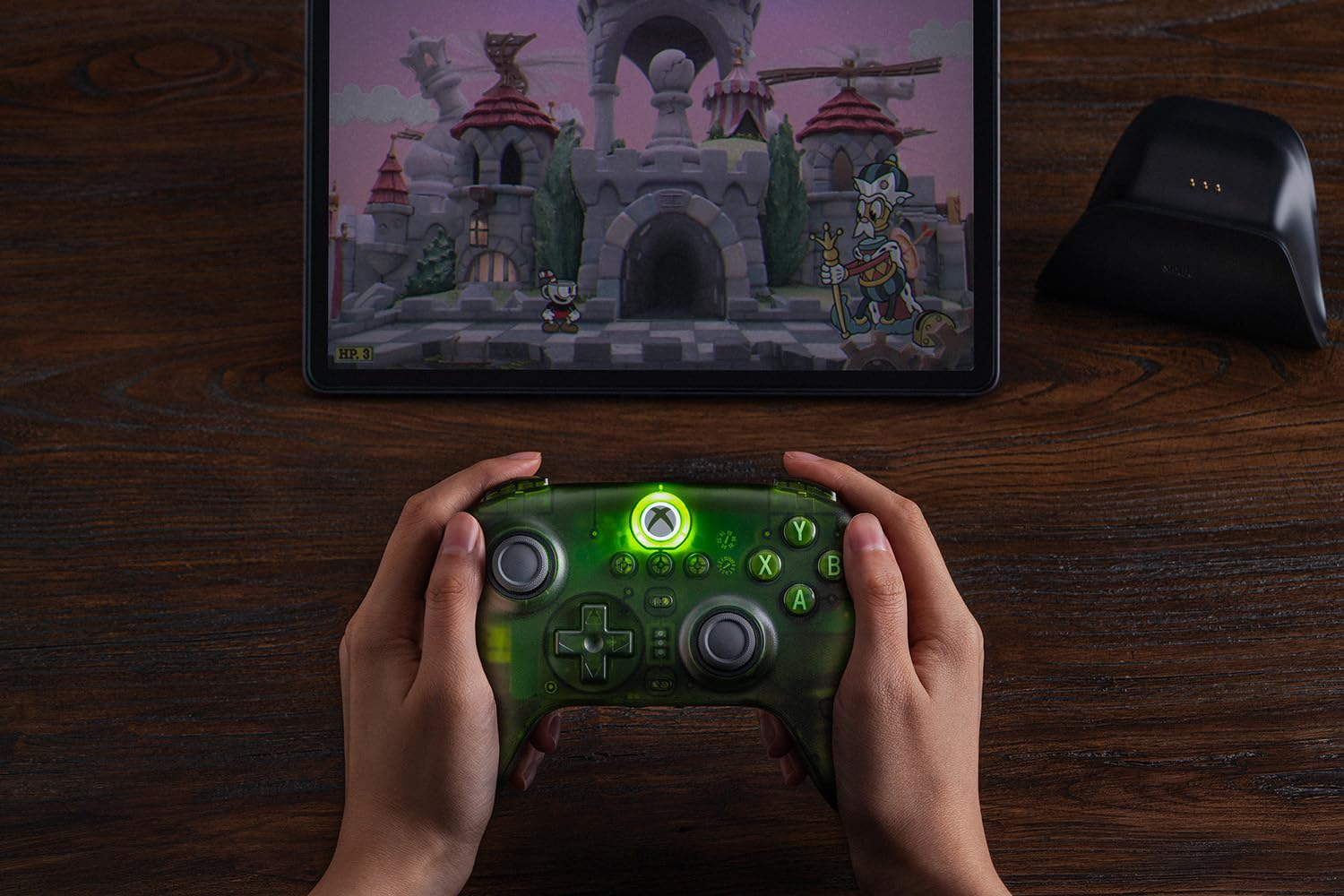 Ręce trzymają zielony kontroler Xbox na drewnianym stole, w tle na tablecie uruchomiona jest gra.