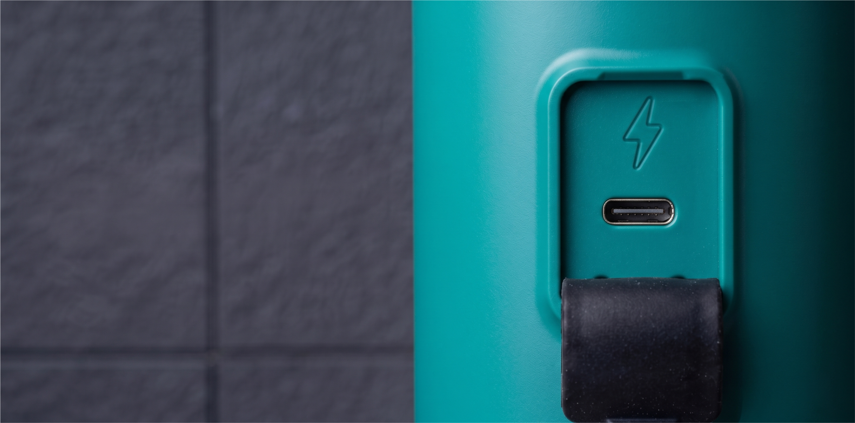 Zbliżenie turkusowego urządzenia z portem USB-C, ikoną błyskawicy i czarną gumową osłoną na tle ciemnej ściany.