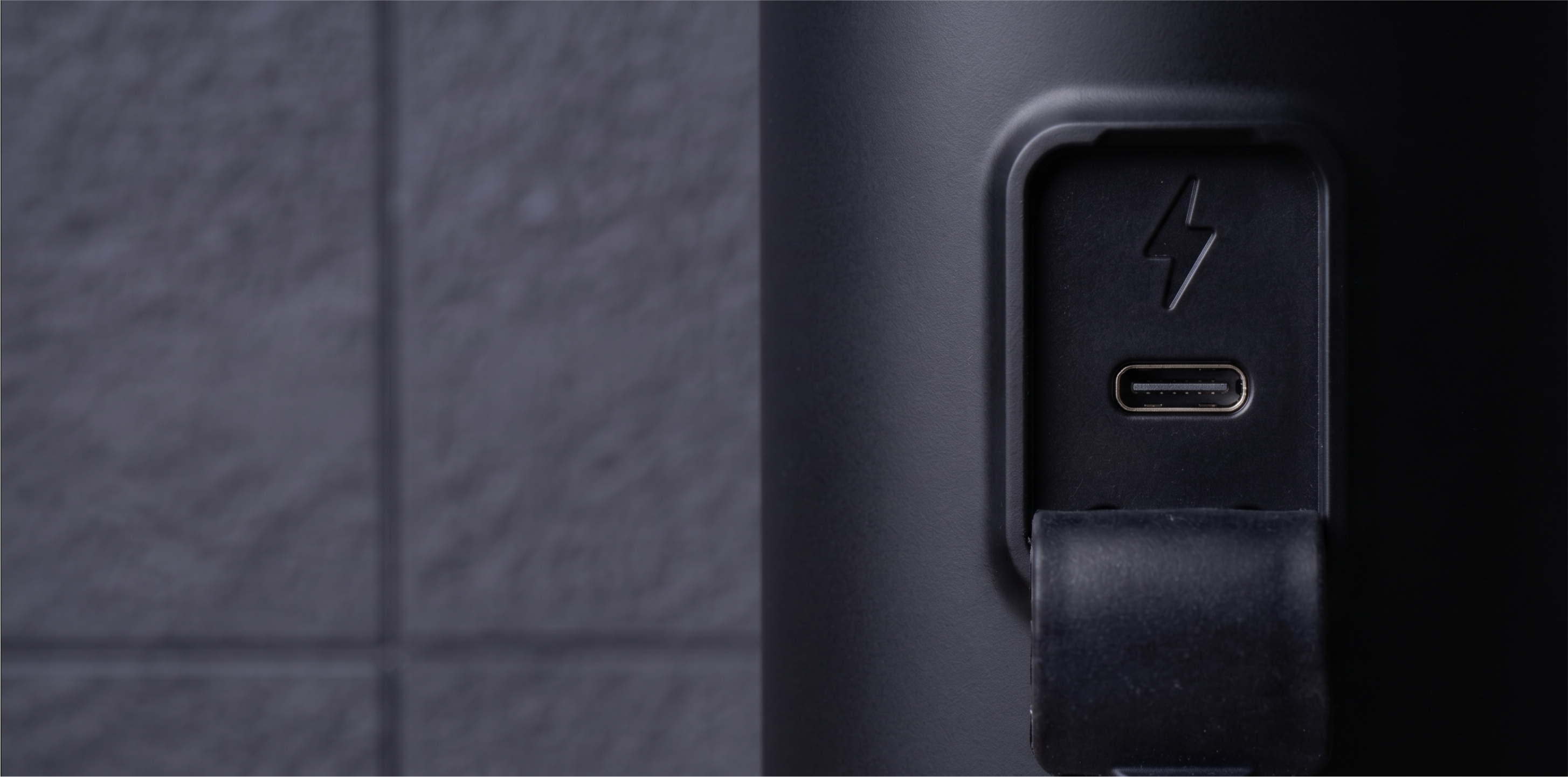 Zbliżenie portu ładowania USB-C z symbolem błyskawicy na czarnym urządzeniu na tle ciemnej ściany.