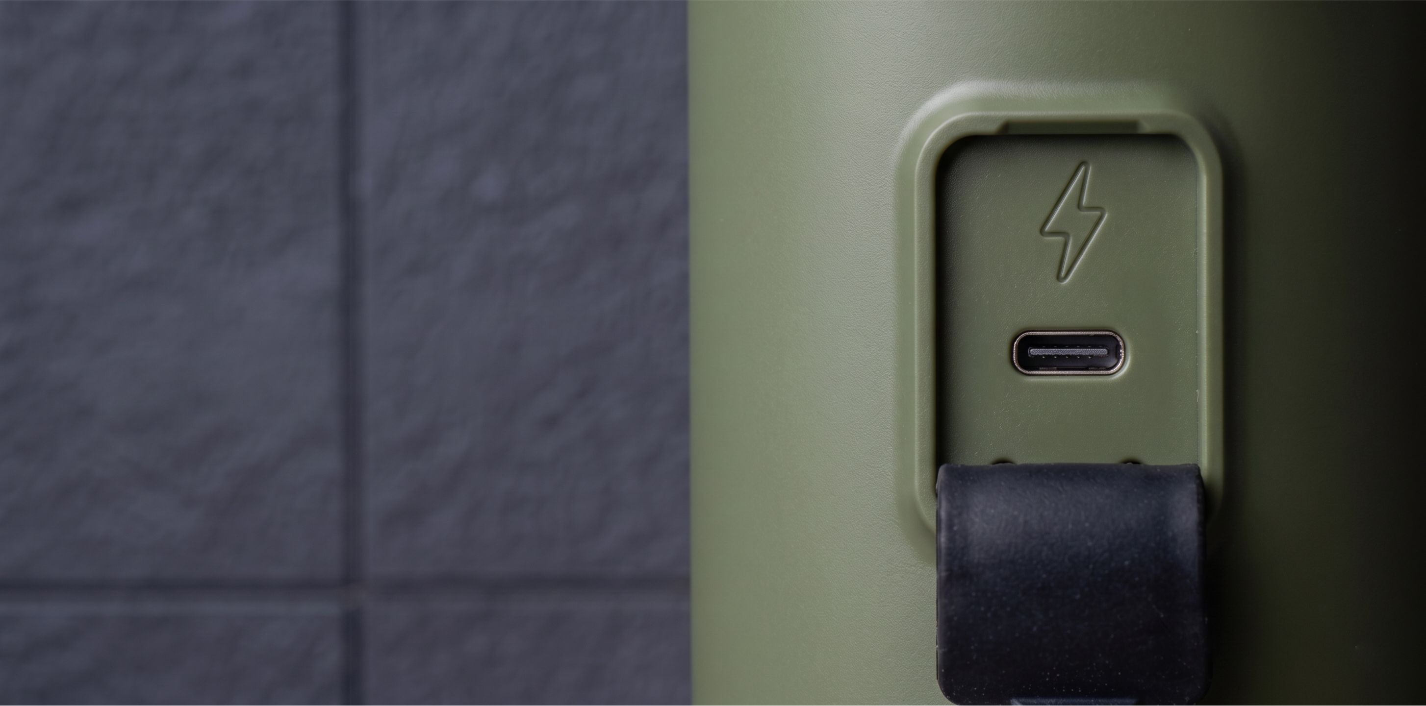 Oliwkowozielone urządzenie z portem USB-C, ikoną błyskawicy i czarną gumową osłoną na tle ciemnej ściany z płytek.