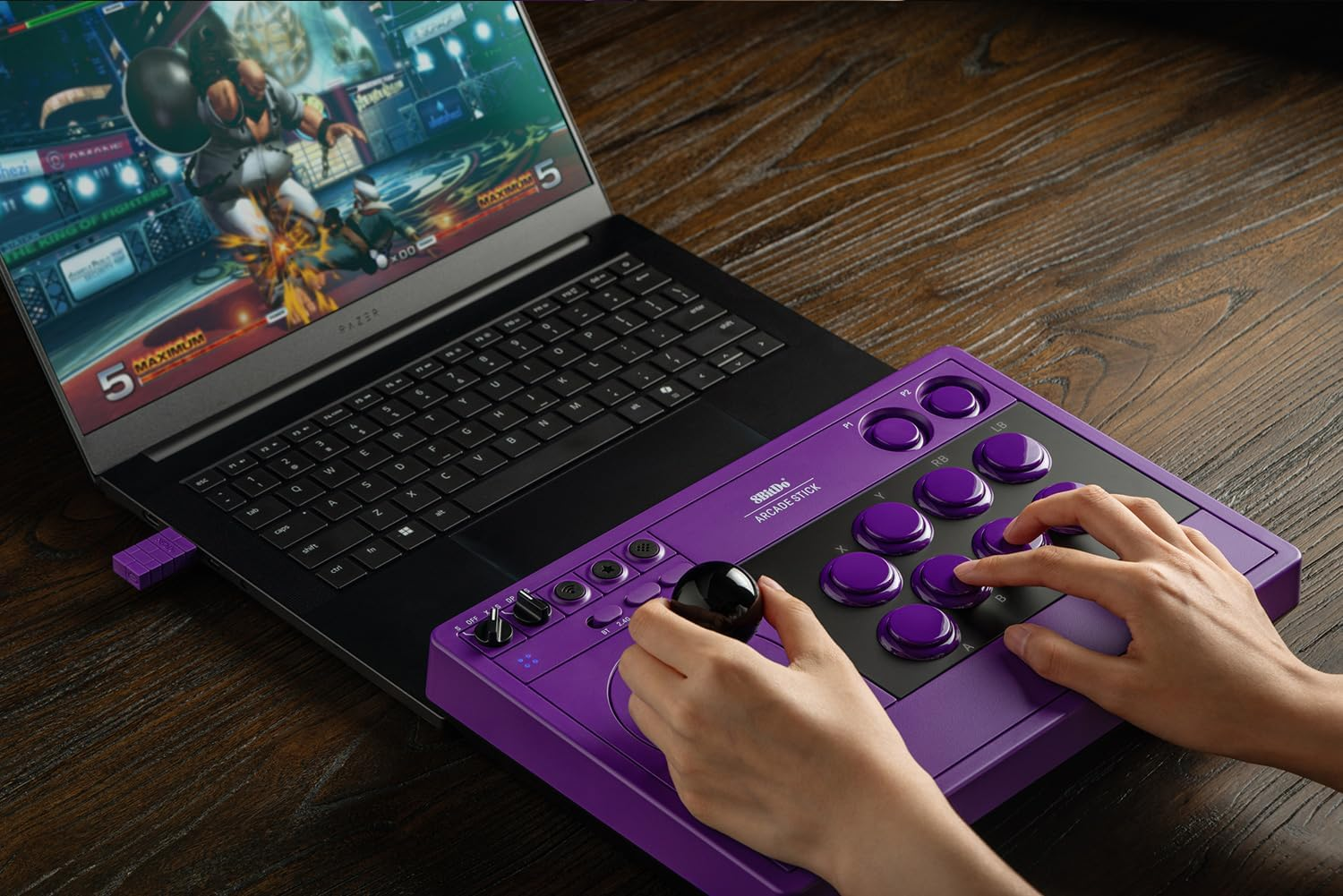 Fioletowy kontroler arcade leży obok laptopa na drewnianym stole, a ktoś na nim gra.