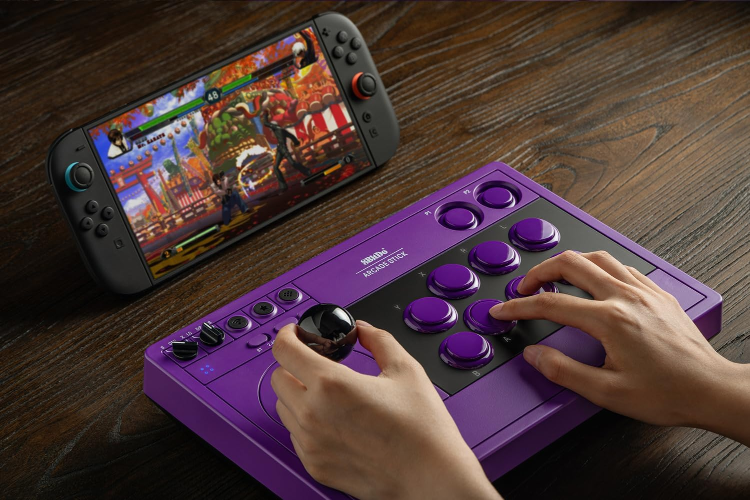 Fioletowy joystick arcade z czarnymi przyciskami leży na drewnianym stole obok konsoli przenośnej.