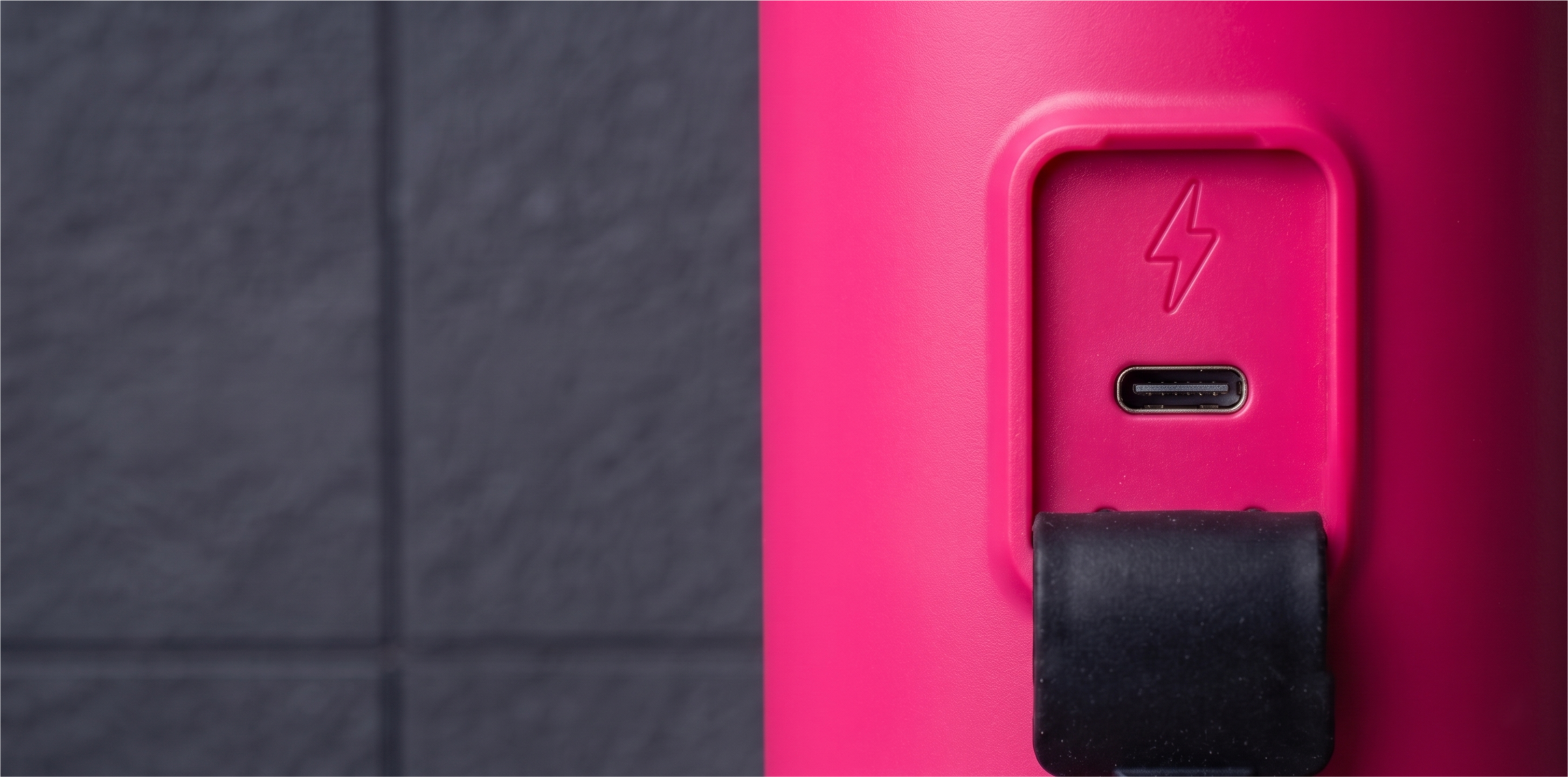 Zbliżenie portu USB-C z symbolem błyskawicy na różowym urządzeniu na tle ciemnej ściany.