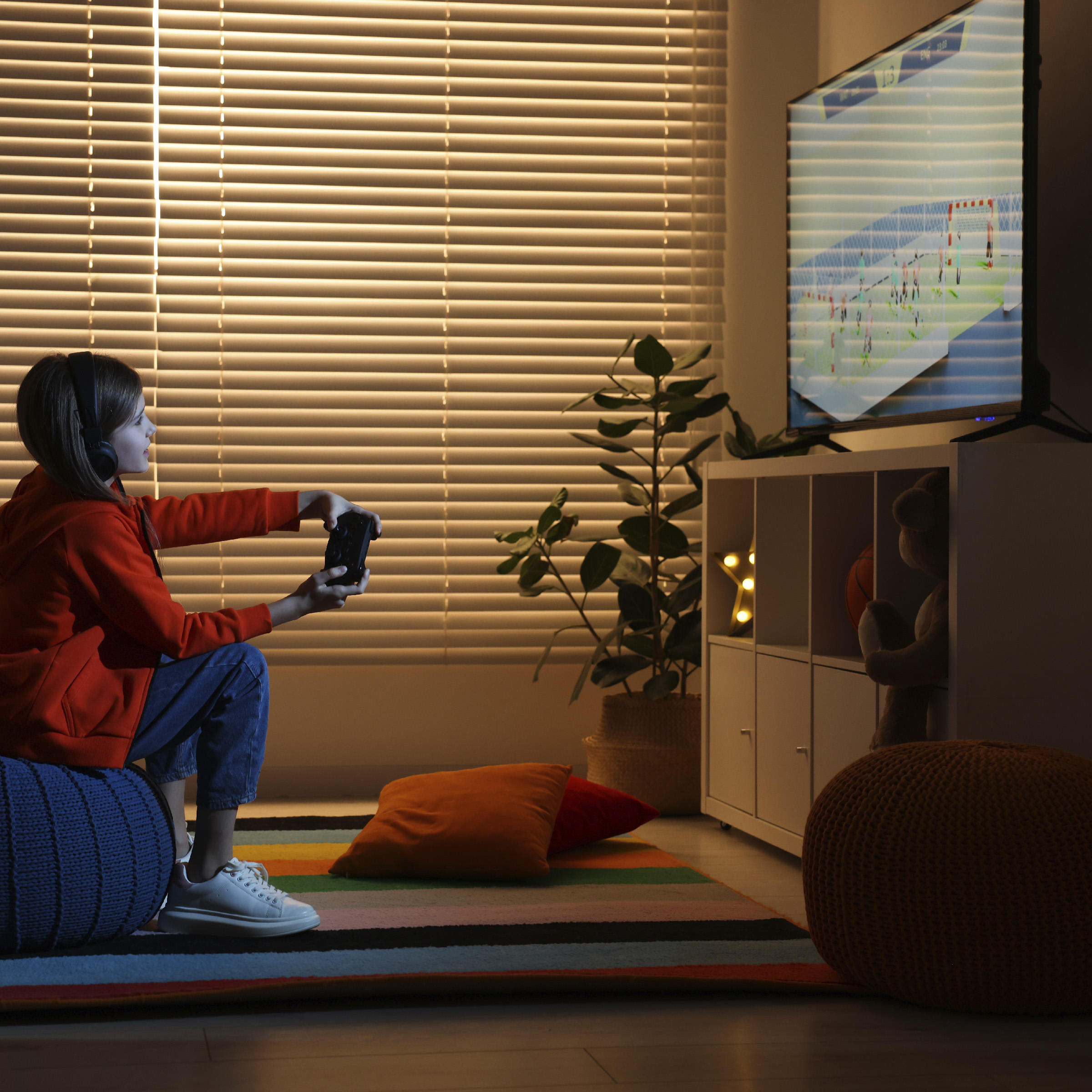 Una niña con suéter rojo y auriculares está sentada en un puf jugando a videojuegos en la televisión.
