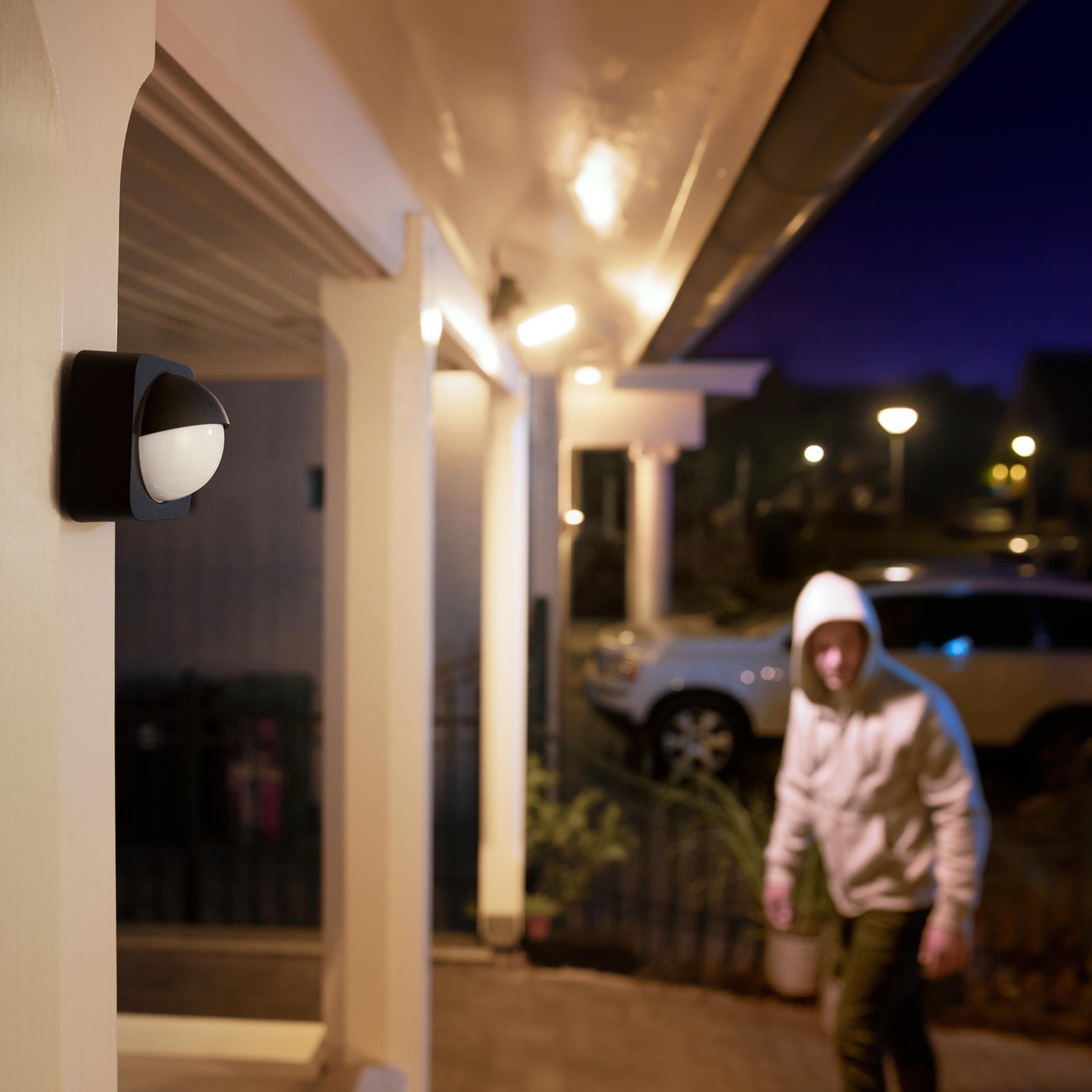 Eine weiß-schwarze Außenlampe leuchtet nachts auf einer Veranda, während eine Person im Kapuzenpullover vorbeigeht.