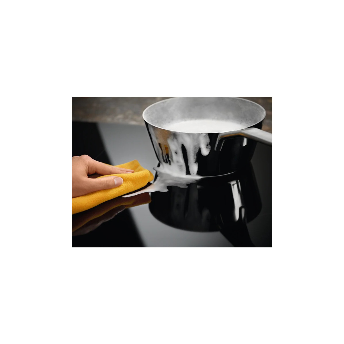 Una mano pulisce la schiuma di sapone bianca da un piano cottura nero accanto a una pentola.