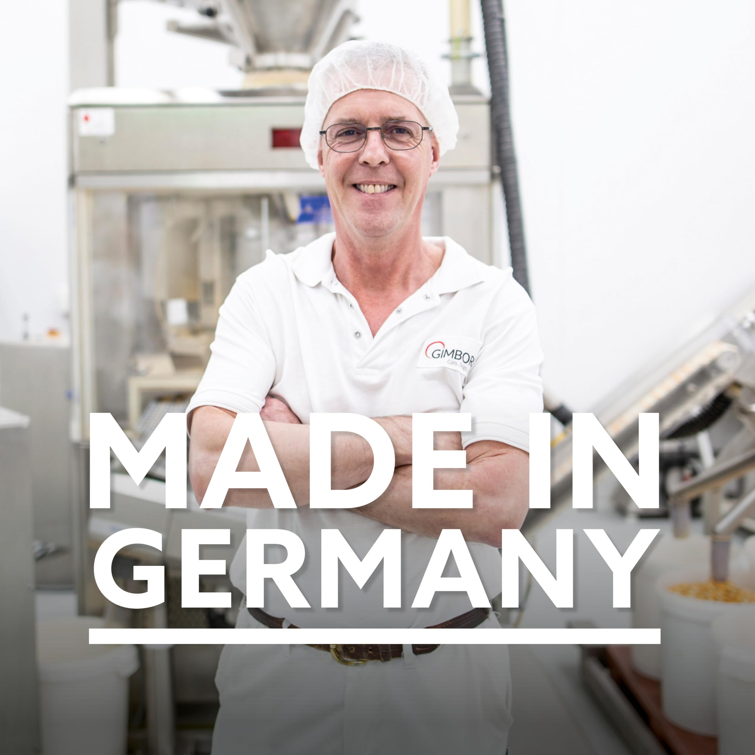Ein Arbeiter in weißer Schutzkleidung und Haarnetz steht mit verschränkten Armen vor einer Maschine in einer Fabrik.
