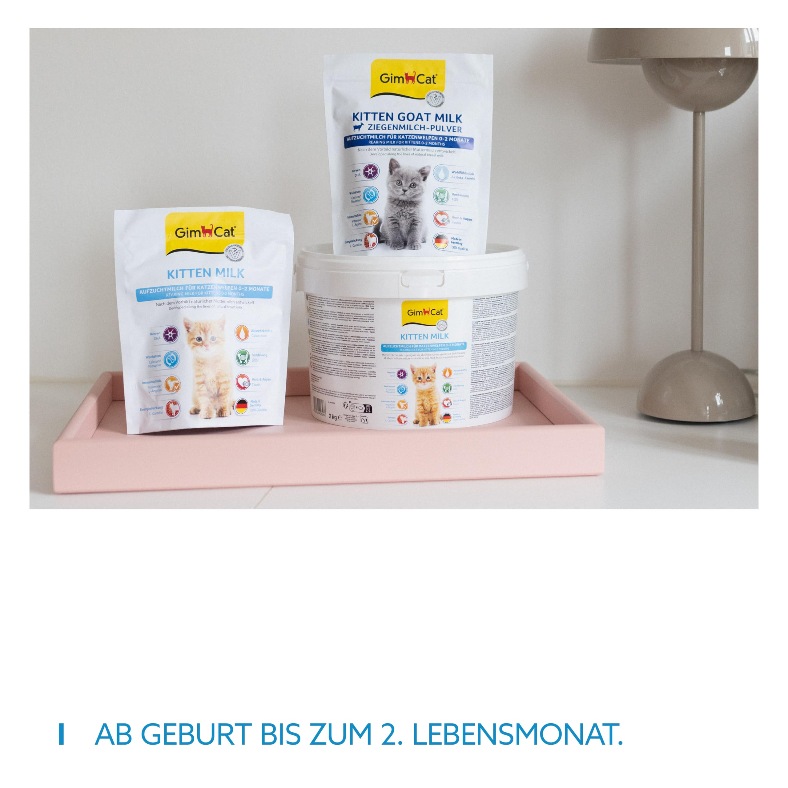 GimCat Katzenmilch-Produkte, darunter zwei Beutel und ein Eimer, auf einem rosa Tablett neben einer Lampe.