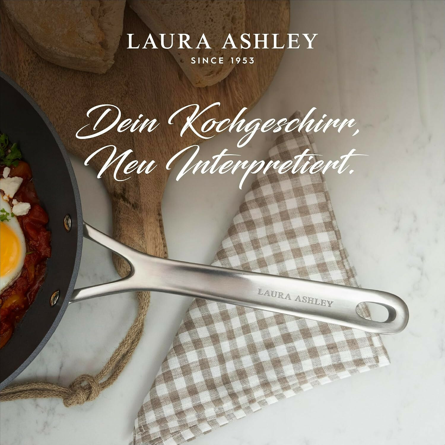 Eine Laura Ashley Bratpfanne mit Spiegelei, Brot und kariertem Geschirrtuch auf hellem Marmor.