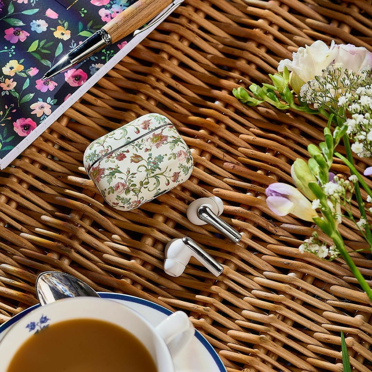 Wicker-Hintergrund mit Kopfhörer-Ladecase, Ohrhörern, floralem Notizbuch, Stift, Blumen und Teetasse.