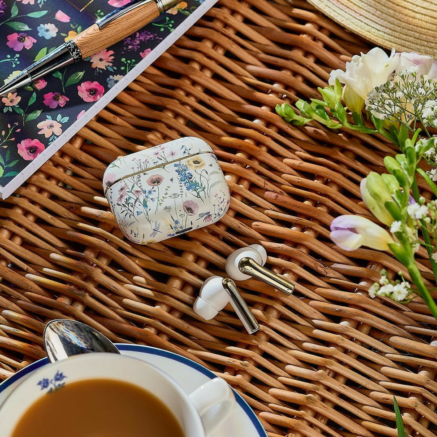 Blumige kabellose Ohrhörer in einem Etui, Notizbuch, Stift, Teetasse und Blumen auf einer Weide.
