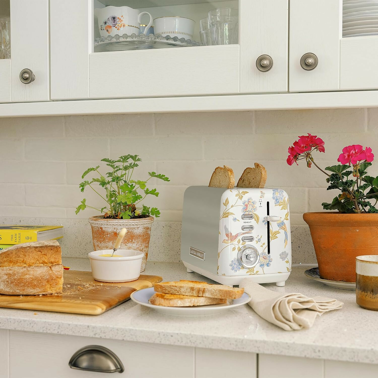 Ein geblümter Toaster mit Toast, frisches Brot, Butter und Pflanzen auf einer weißen Küchentheke.
