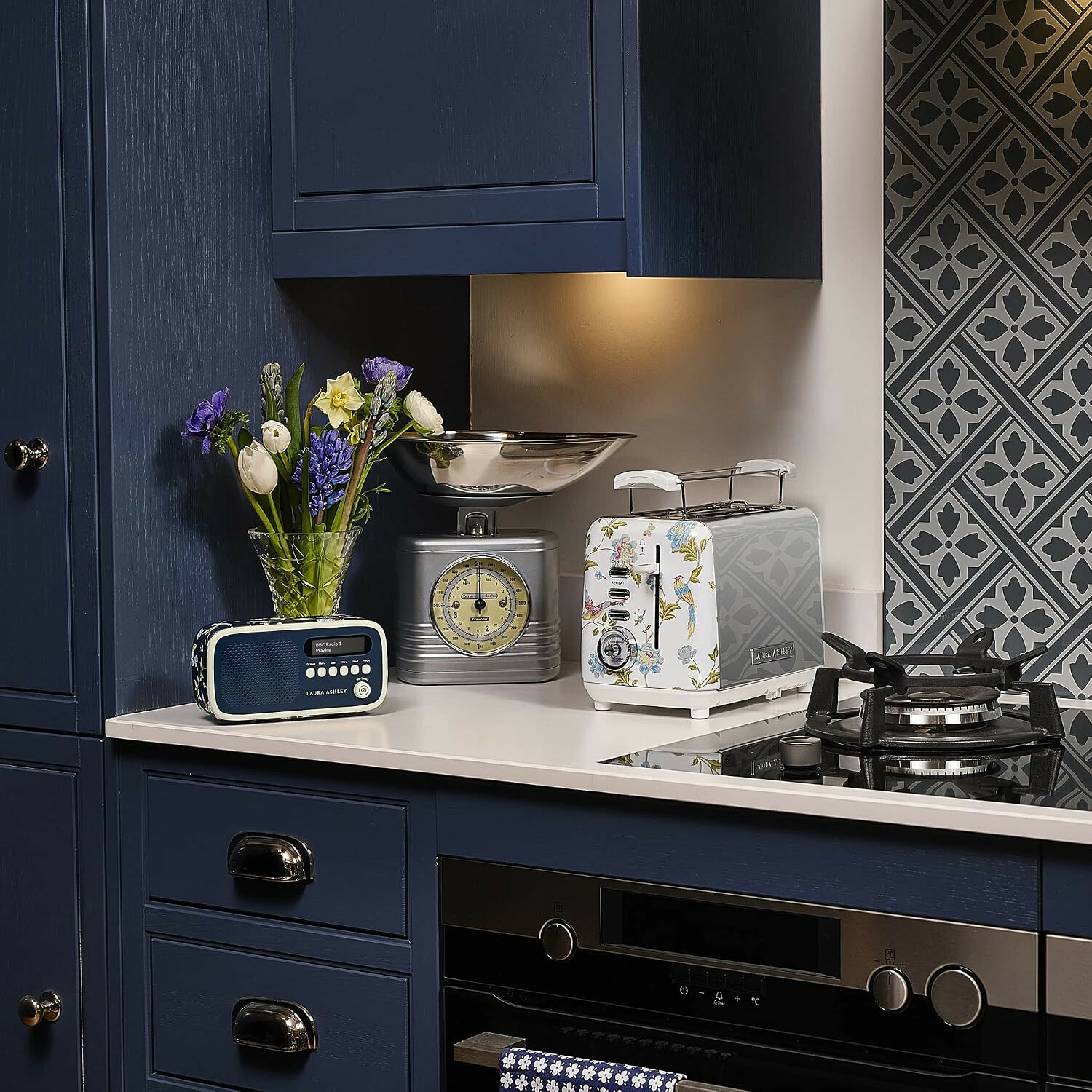 Blaue Küche mit weißer Arbeitsplatte, gemusterter Toaster, Digitalradio, Küchenwaage und Blumen in einer Vase.