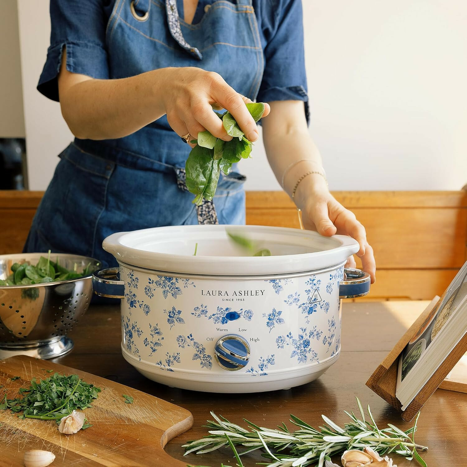 Eine Frau im blauen Denim-Apron fügt Spinat in einen weiß-blauen Laura-Ashley-Slow-Cooker auf Holztisch.