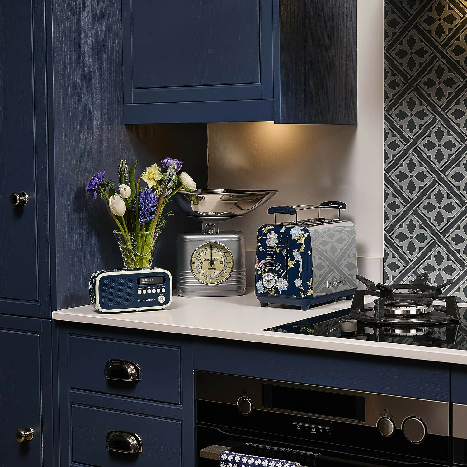 Blaue Küche mit Blumen, Radio, Waage und Toaster auf weißer Arbeitsplatte vor gemusterten Fliesen.