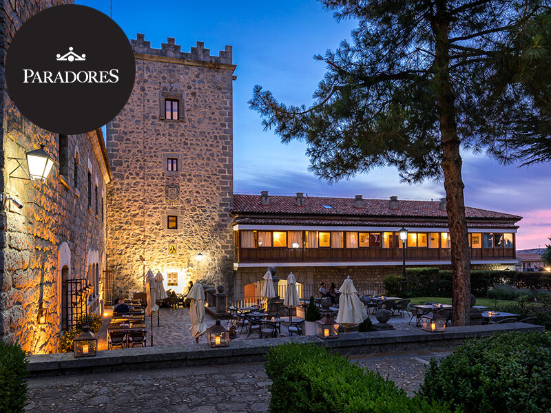 Un castillo de piedra con patio iluminado, mesas y grandes pinos al atardecer.