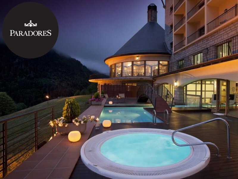 Jacuzzi iluminado en primer plano con un hotel y piscina al fondo al atardecer.