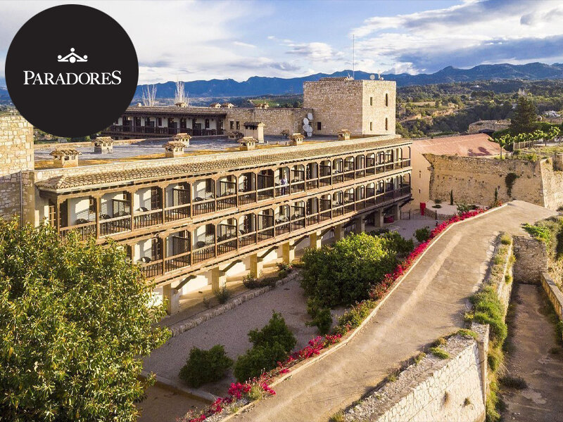 Vista aérea de un edificio histórico de piedra con balcones de madera y jardines ante las montañas.