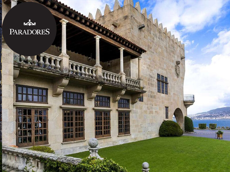 Castillo de piedra histórico con balcón, patio de césped y vista al mar al fondo.