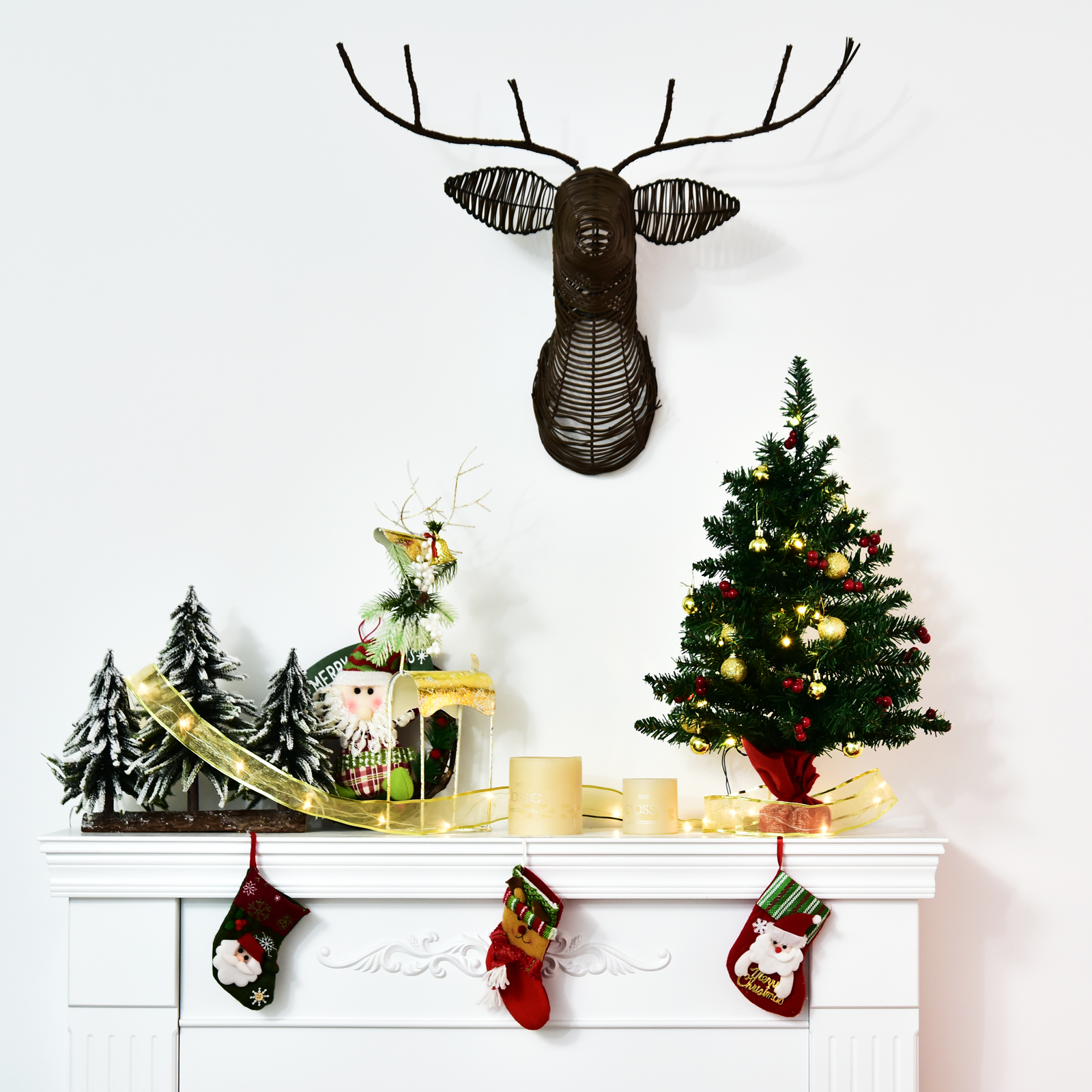 Una repisa de chimenea blanca decorada para Navidad con calcetines, árboles pequeños, un árbol iluminado y una cabeza de ciervo de alambre en la pared.