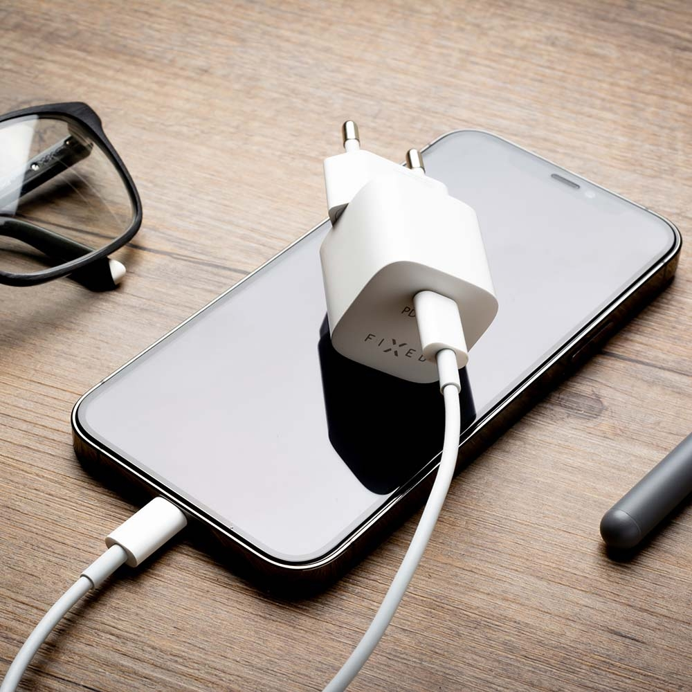 Ein Smartphone auf einem Holztisch, verbunden mit weißem Ladegerät und Kabel, Brille daneben.
