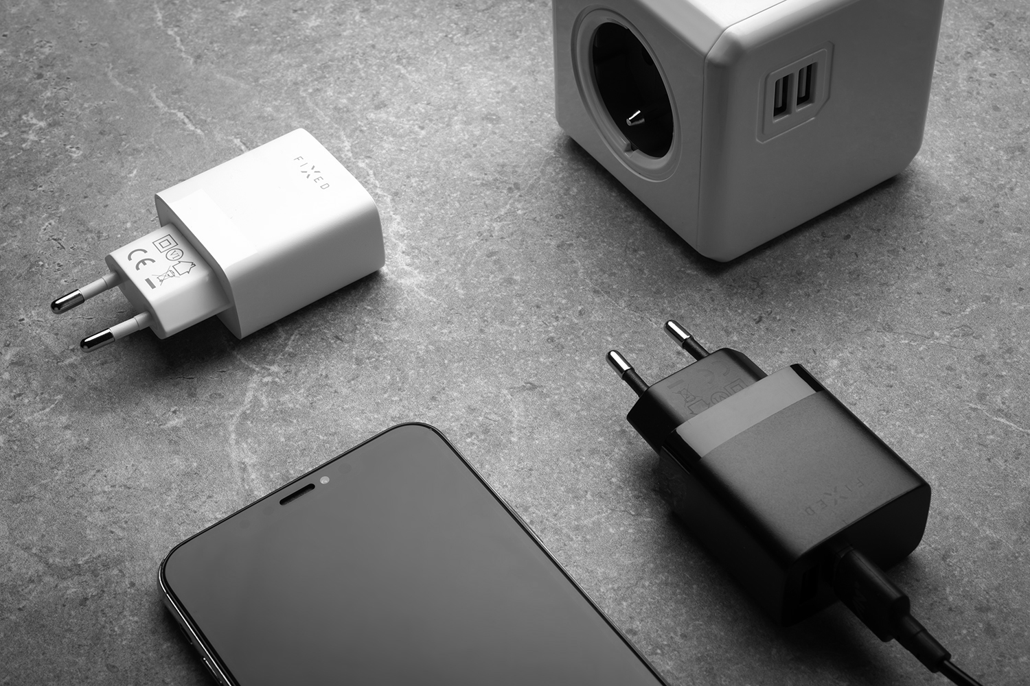 Ein Smartphone, zwei Wandladegeräte und ein Steckdosenwürfel liegen auf einer grauen Oberfläche.