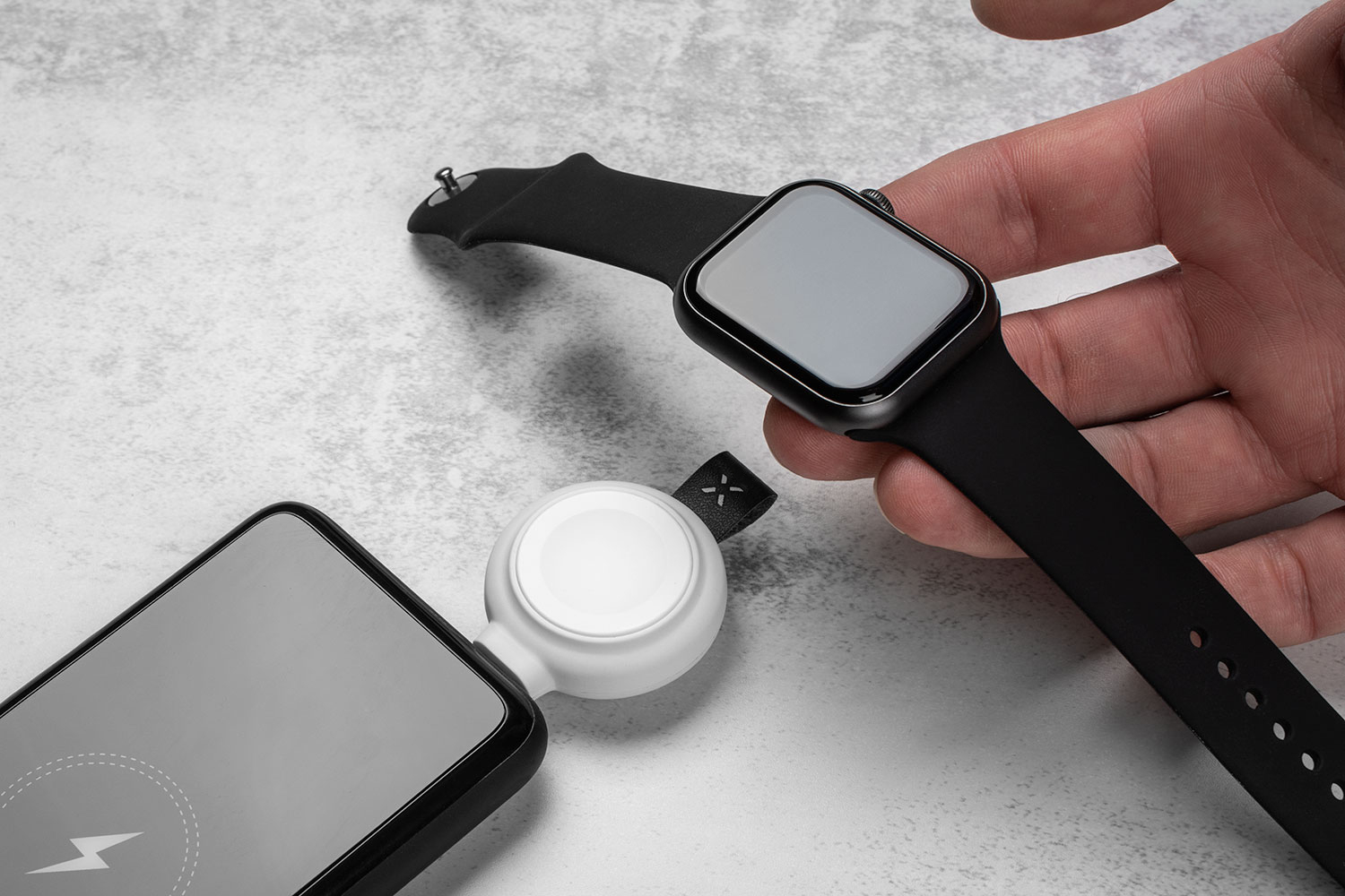 Eine Hand hält eine schwarze Smartwatch über ein weißes Ladegerät, das mit einem Handy verbunden ist.