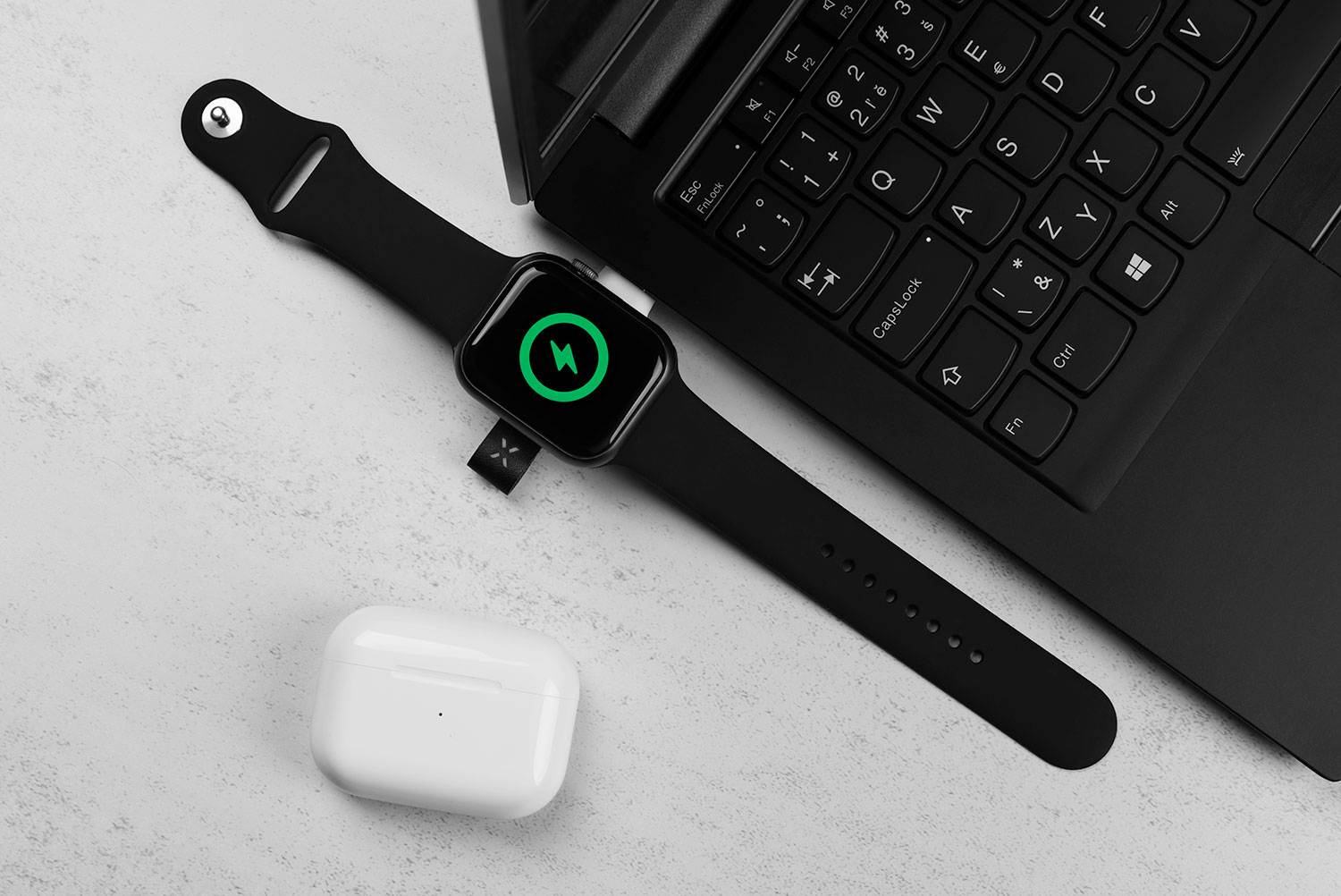 Eine schwarze Smartwatch mit grünem Ladesymbol liegt neben einem Laptop und weißen Ohrhörern auf einem Tisch.