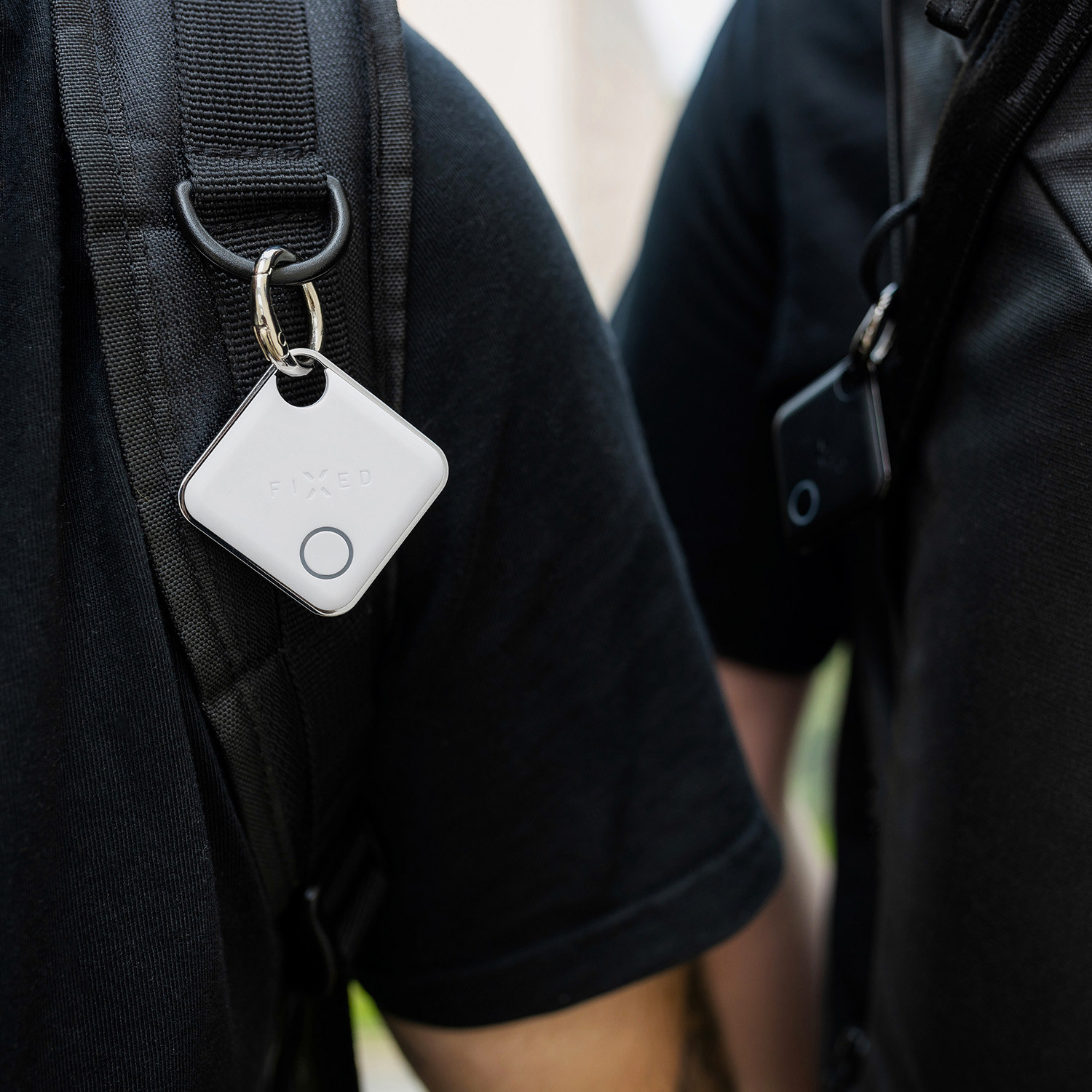 Ein weißer, quadratischer FIXED Smart-Tracker hängt am Rucksackriemen einer Person in schwarzem Hemd.