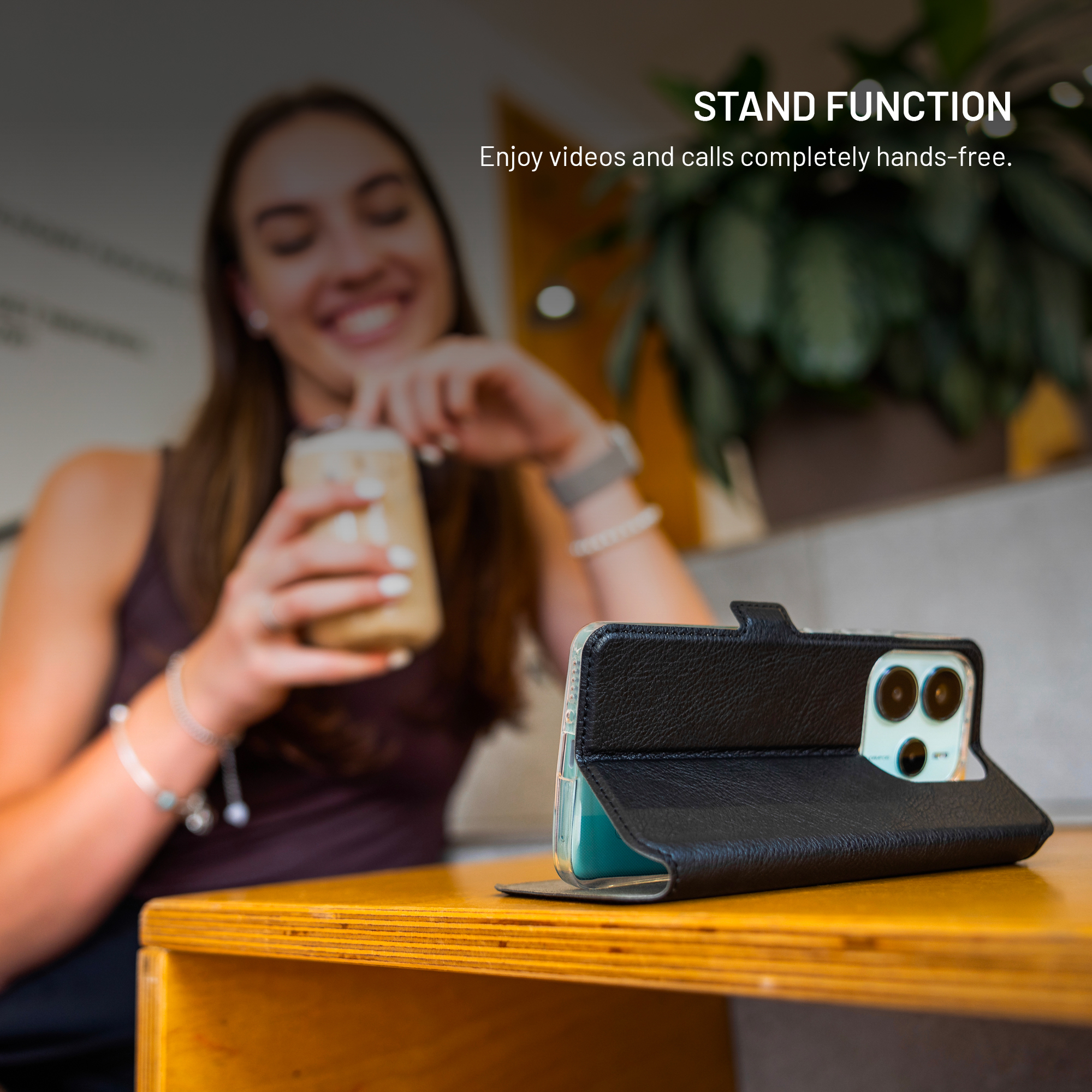 Ein Smartphone in einer schwarzen Ledertasche steht auf einem Holztisch, im Hintergrund sitzt eine lächelnde Frau.