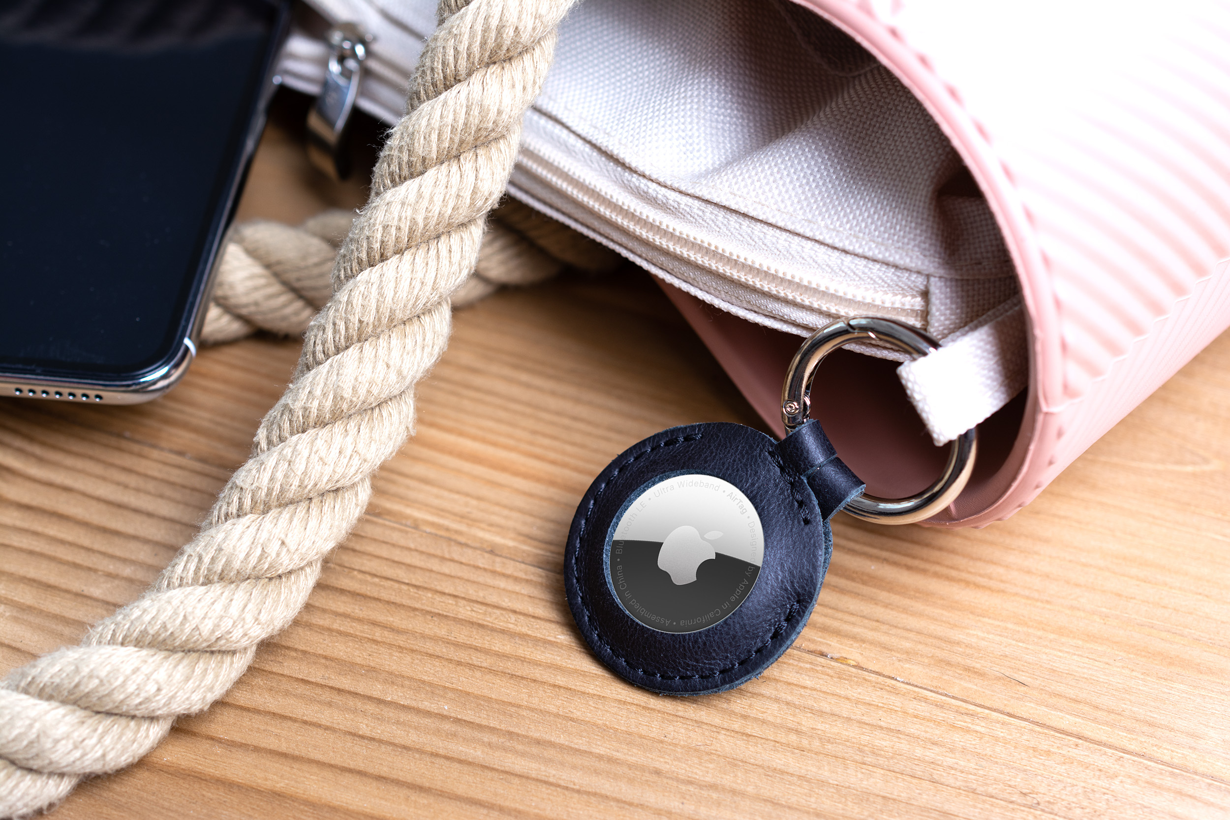Ein Apple AirTag in schwarzem Lederetui, an einer rosa Handtasche mit Kordel auf Holztisch befestigt.