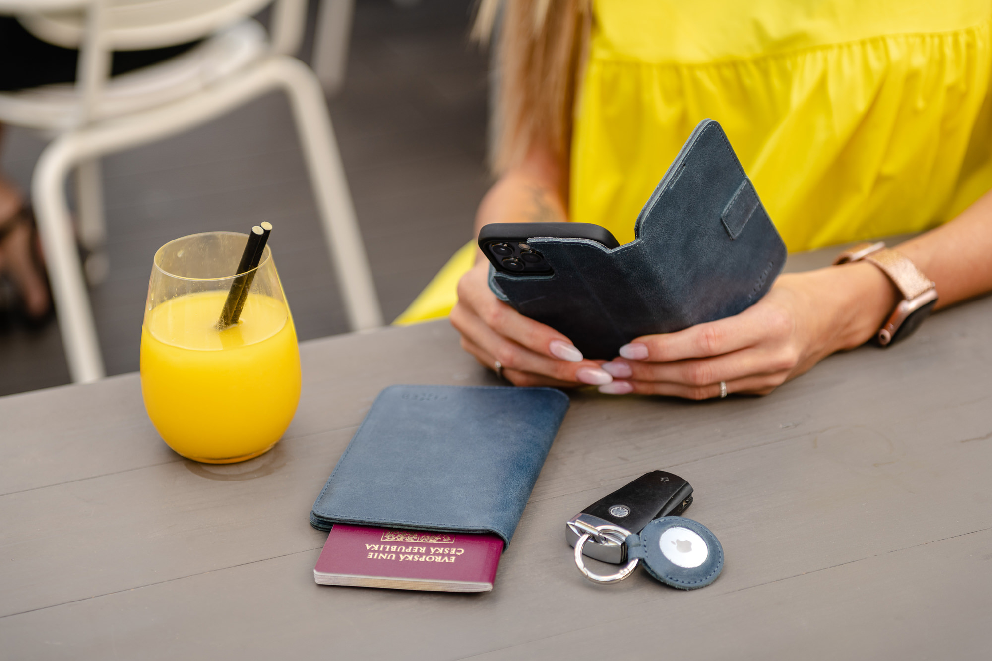 Eine Frau in Gelb sitzt am Tisch mit Orangensaft, einem blauen Pass und Schlüsseln, während sie ihr Telefon hält.