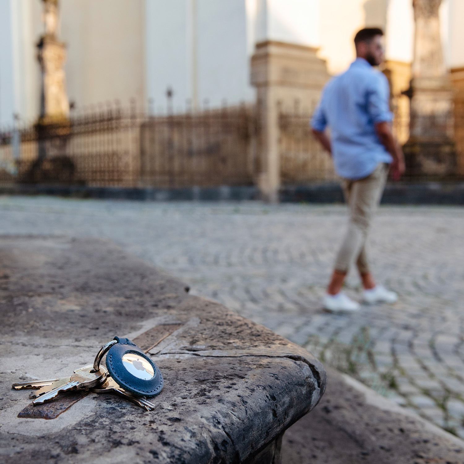 Ein Schlüsselbund mit blauem Leder-Tracker liegt im Freien auf Stein, im Hintergrund geht ein Mann.
