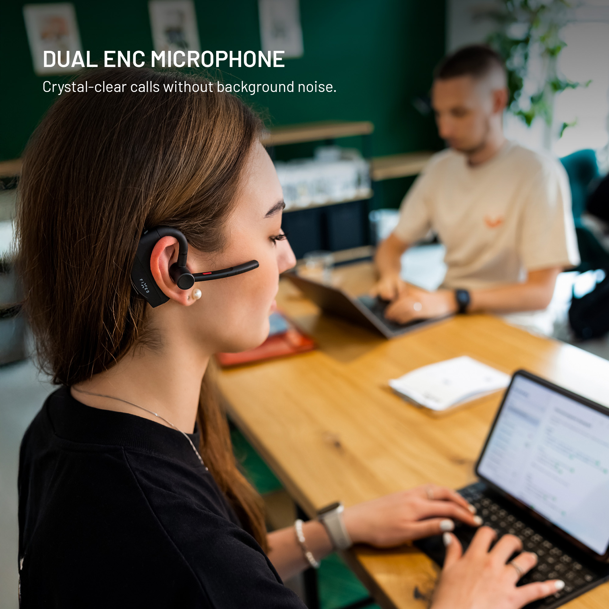 Eine Frau mit einem schwarzen Headset tippt an einem Tisch auf ein Tablet, während ein Mann im Hintergrund arbeitet.