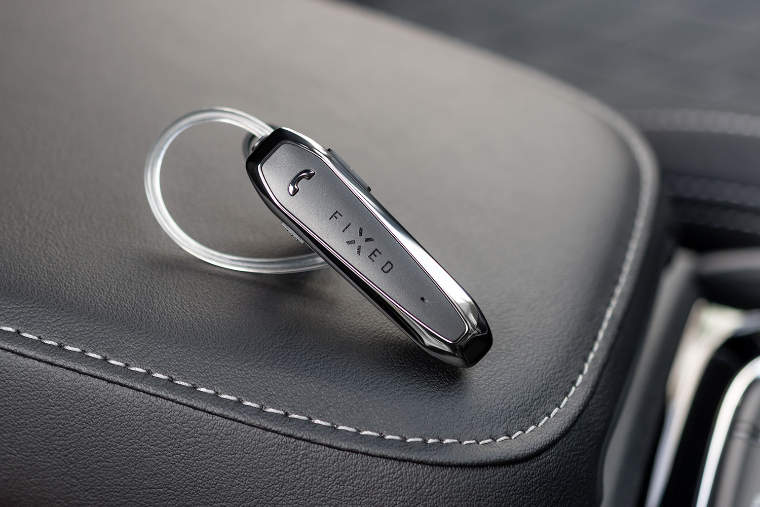 Ein schwarzes FIXED Bluetooth-Headset mit transparentem Ohrbügel liegt auf einer dunklen Lederoberfläche.