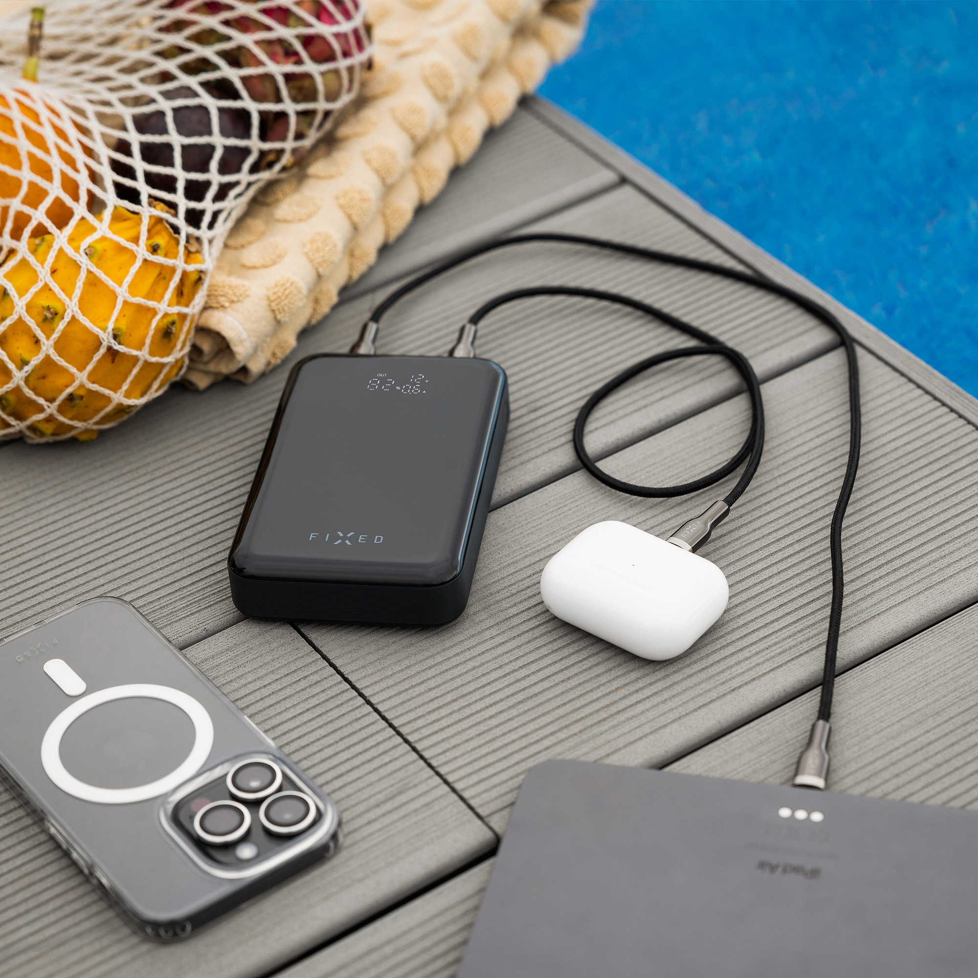 Eine schwarze Powerbank lädt Kopfhörer und ein Tablet auf einer Holzbühne neben Obst und einem Handtuch.