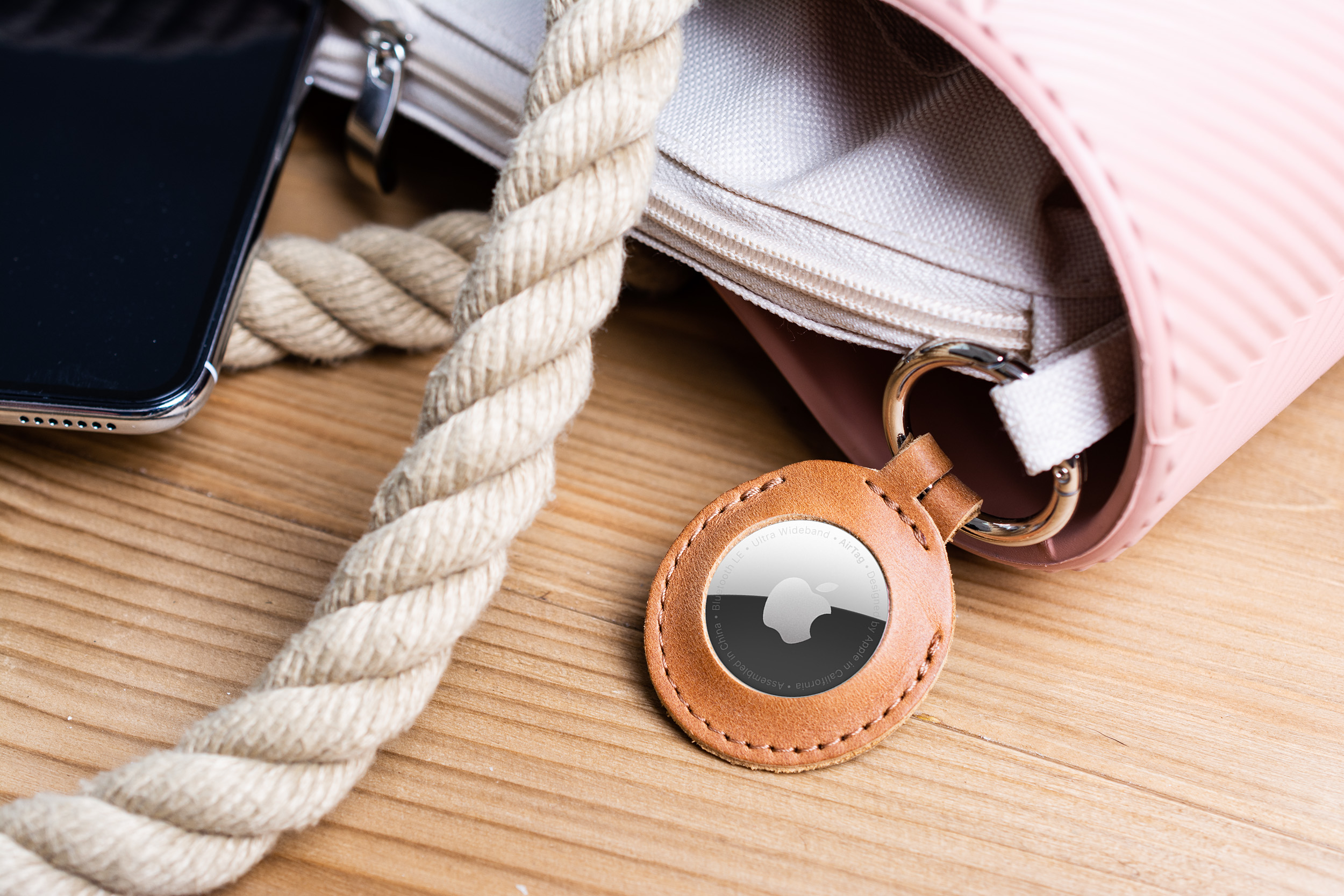 Ein Apple AirTag in einem Lederanhänger hängt an einer rosa Handtasche, neben einem Seil und einem Smartphone auf Holz.