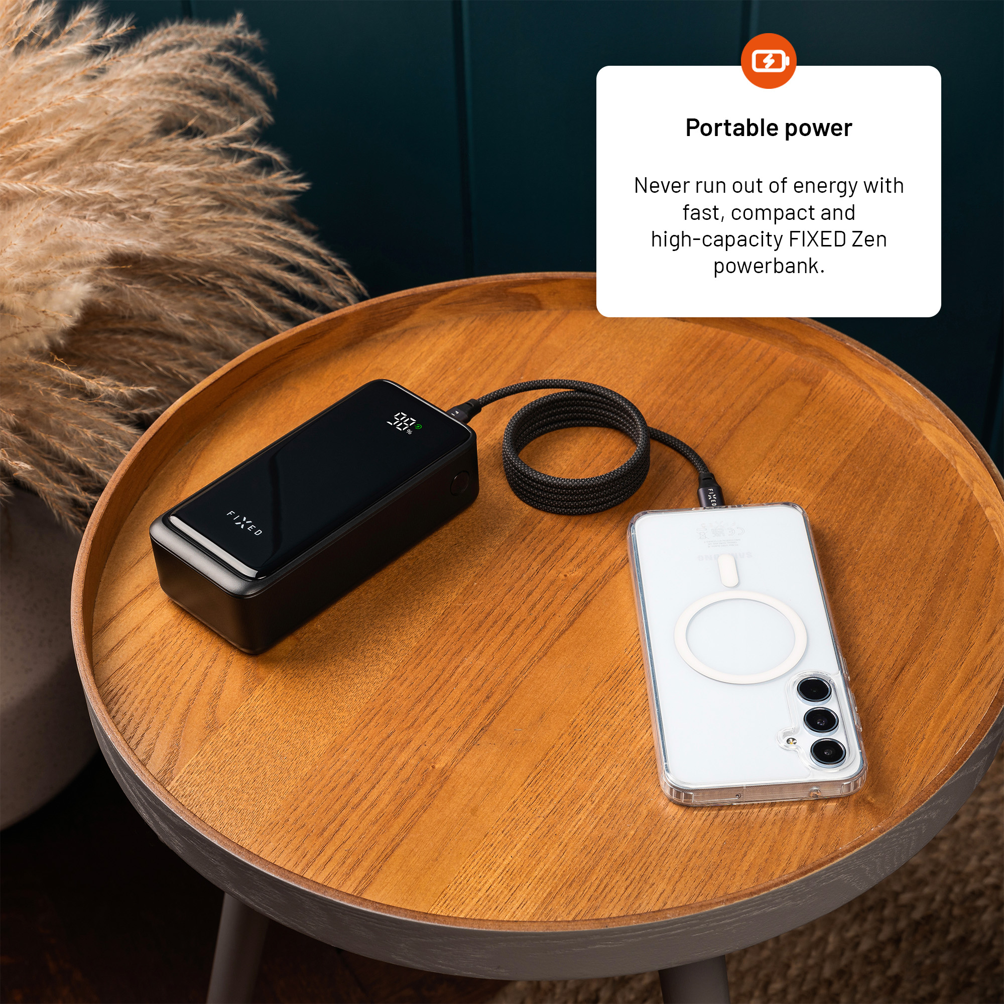 Eine schwarze Powerbank lädt ein Smartphone auf einem Holztisch neben Pampasgras.