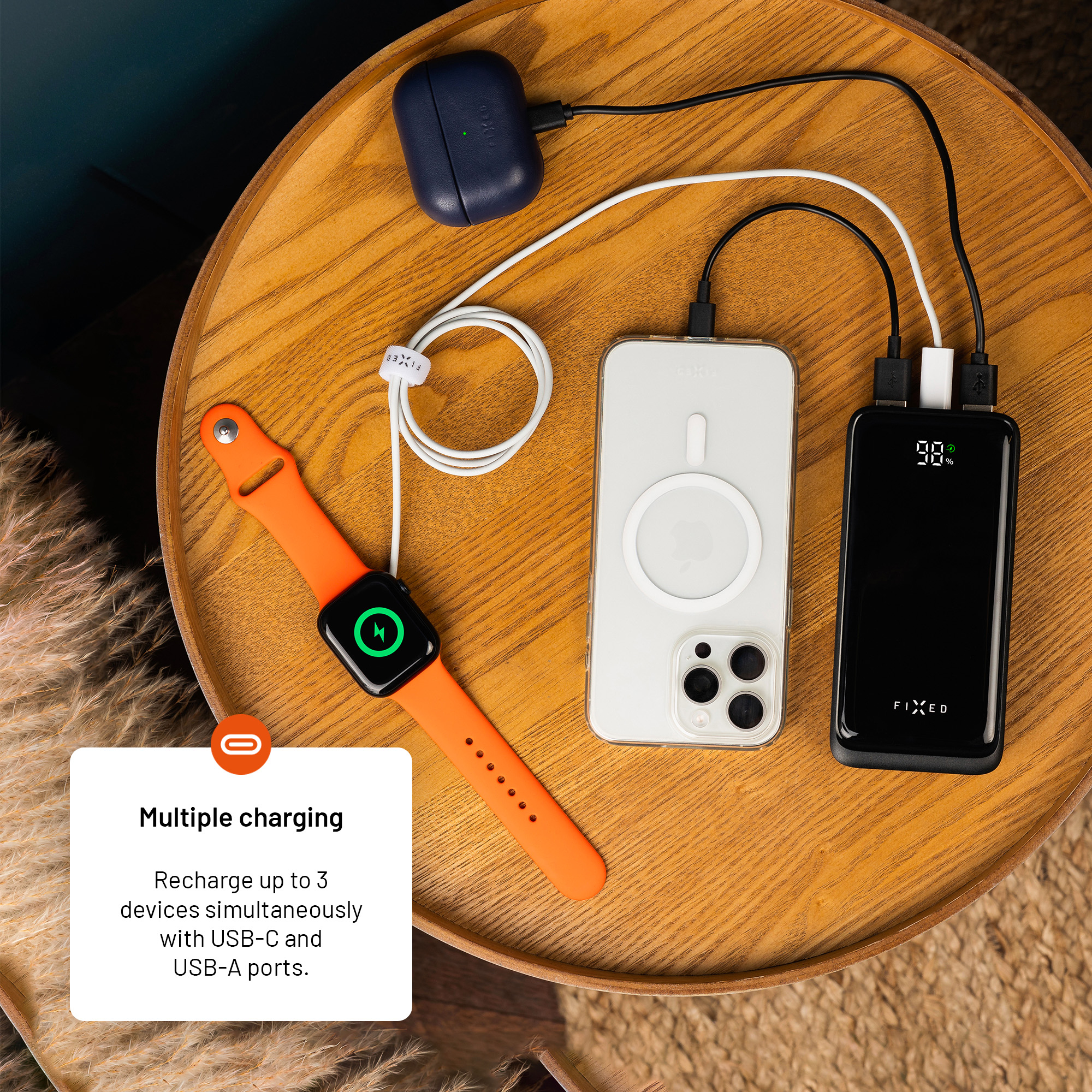 Eine Powerbank lädt gleichzeitig eine Smartwatch, ein Smartphone und Ohrhörer auf einem runden Holztisch.