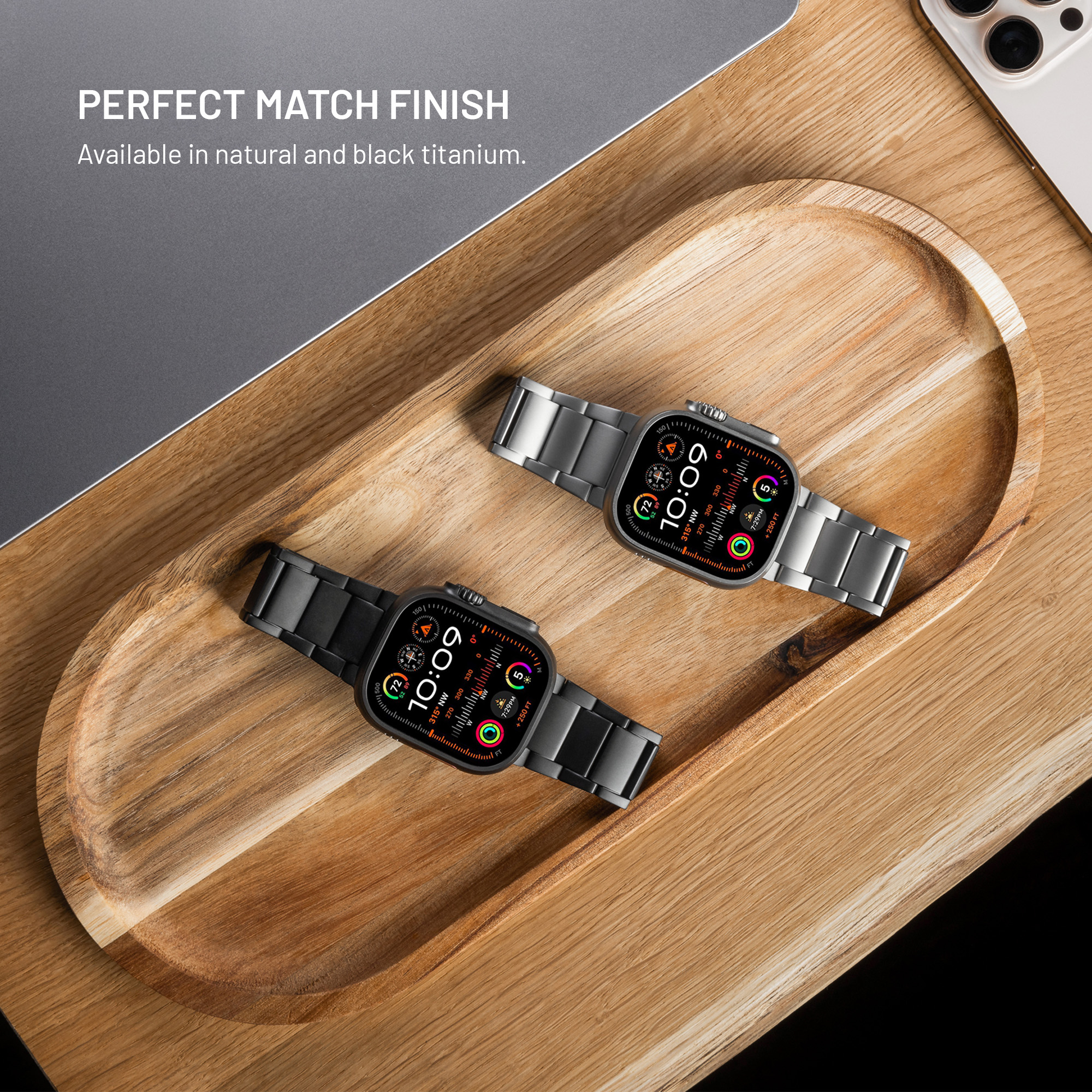 Zwei Smartwatches in natürlichem und schwarzem Titan liegen auf einem Holztablett neben einem Laptop.