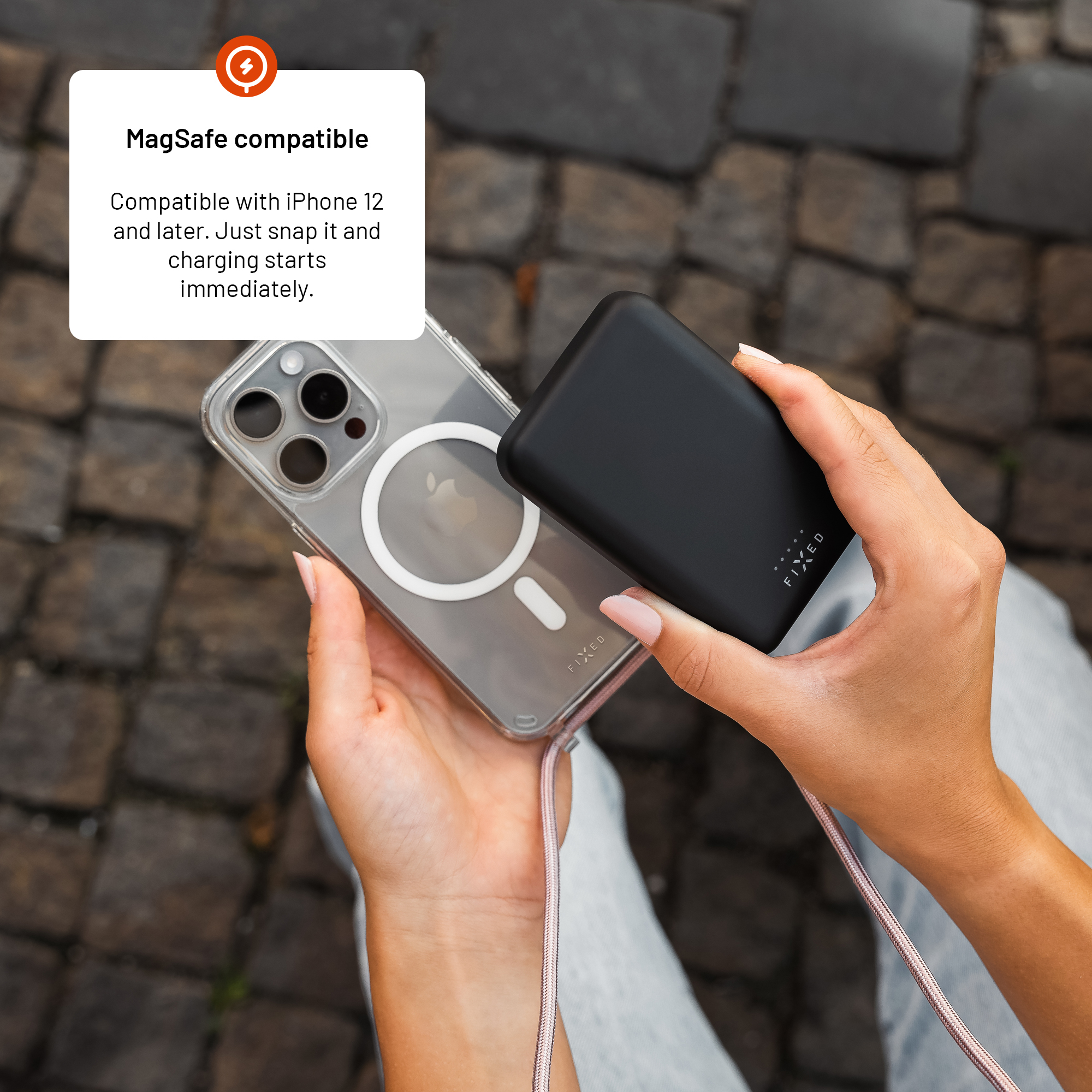 Person hält ein iPhone mit Hülle und ein schwarzes MagSafe-Powerbank vor Pflasterstein.
