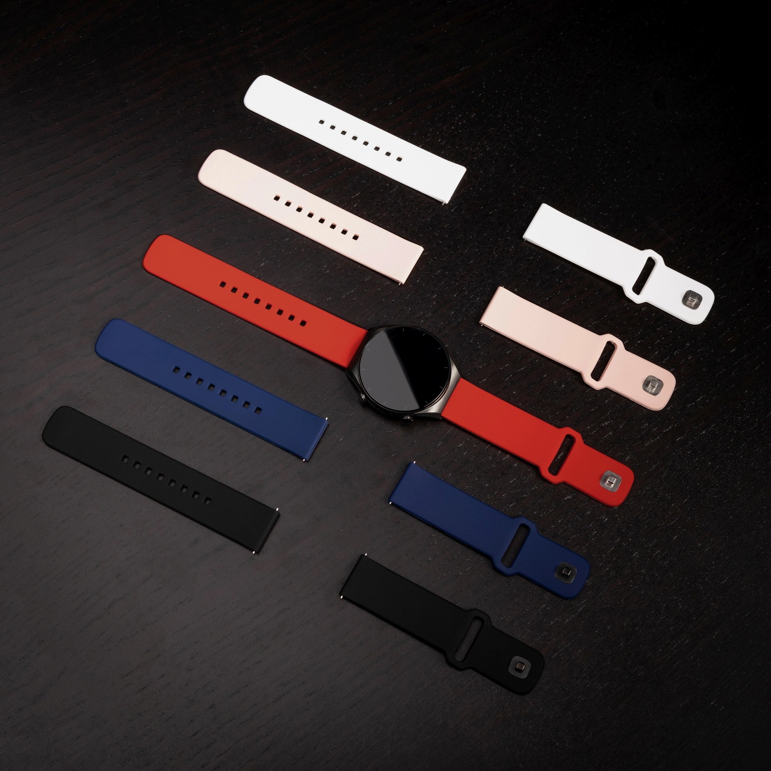 Eine schwarze Smartwatch mit rotem Armband ist von austauschbaren Armbändern in Weiß, Rosa, Blau und Schwarz umgeben.