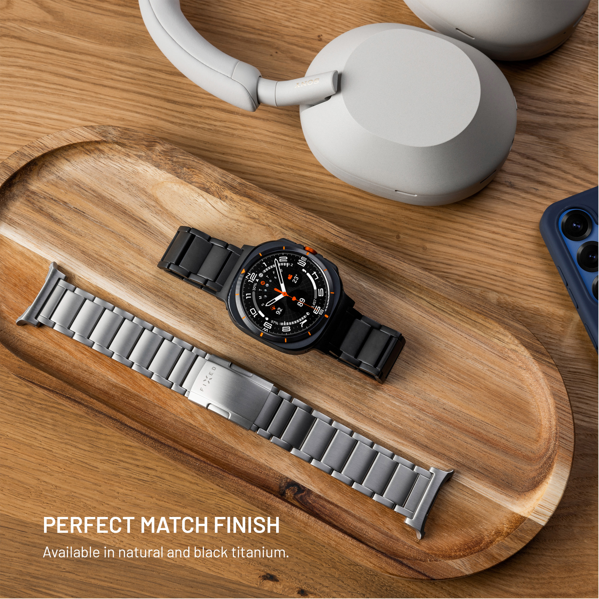 Eine schwarze Smartwatch und ein silbernes Metallarmband auf einem Holztablett mit Kopfhörern.