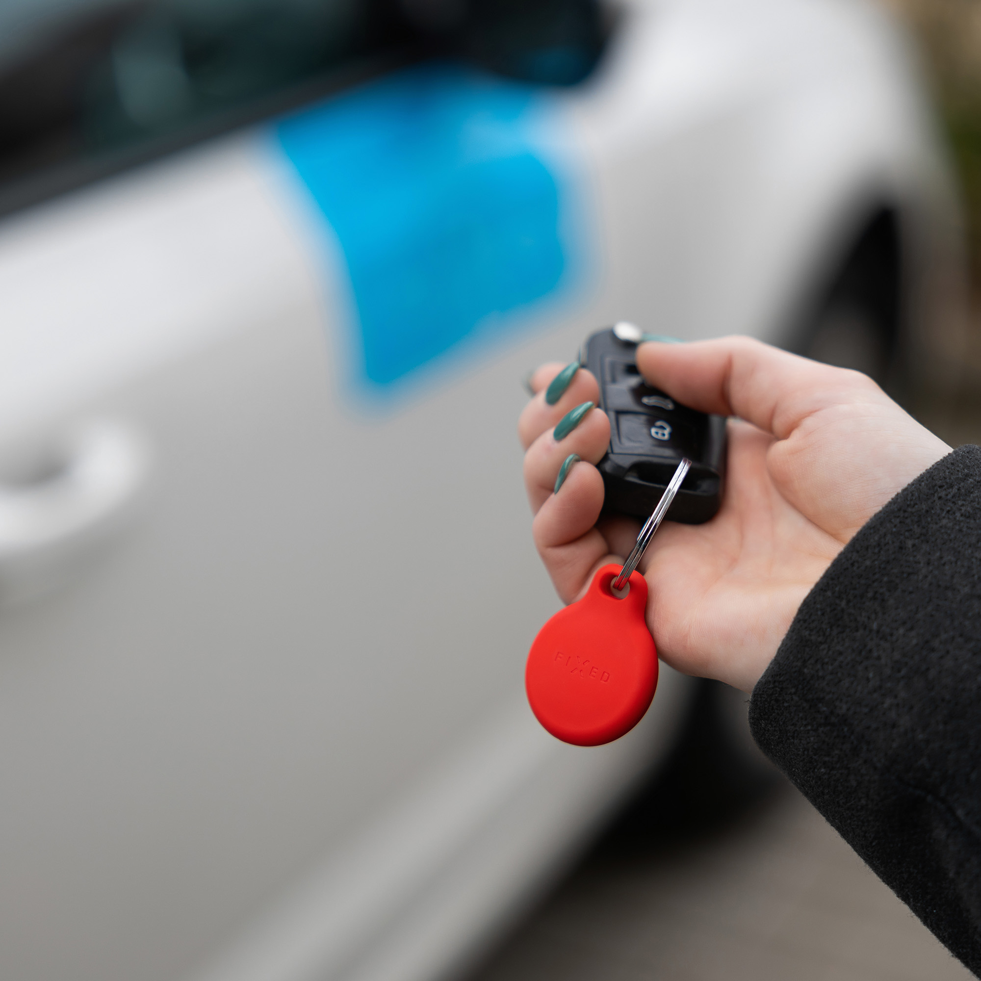Eine Hand hält einen Autoschlüssel mit rotem Anhänger vor ein weißes Auto mit blauem Schild.