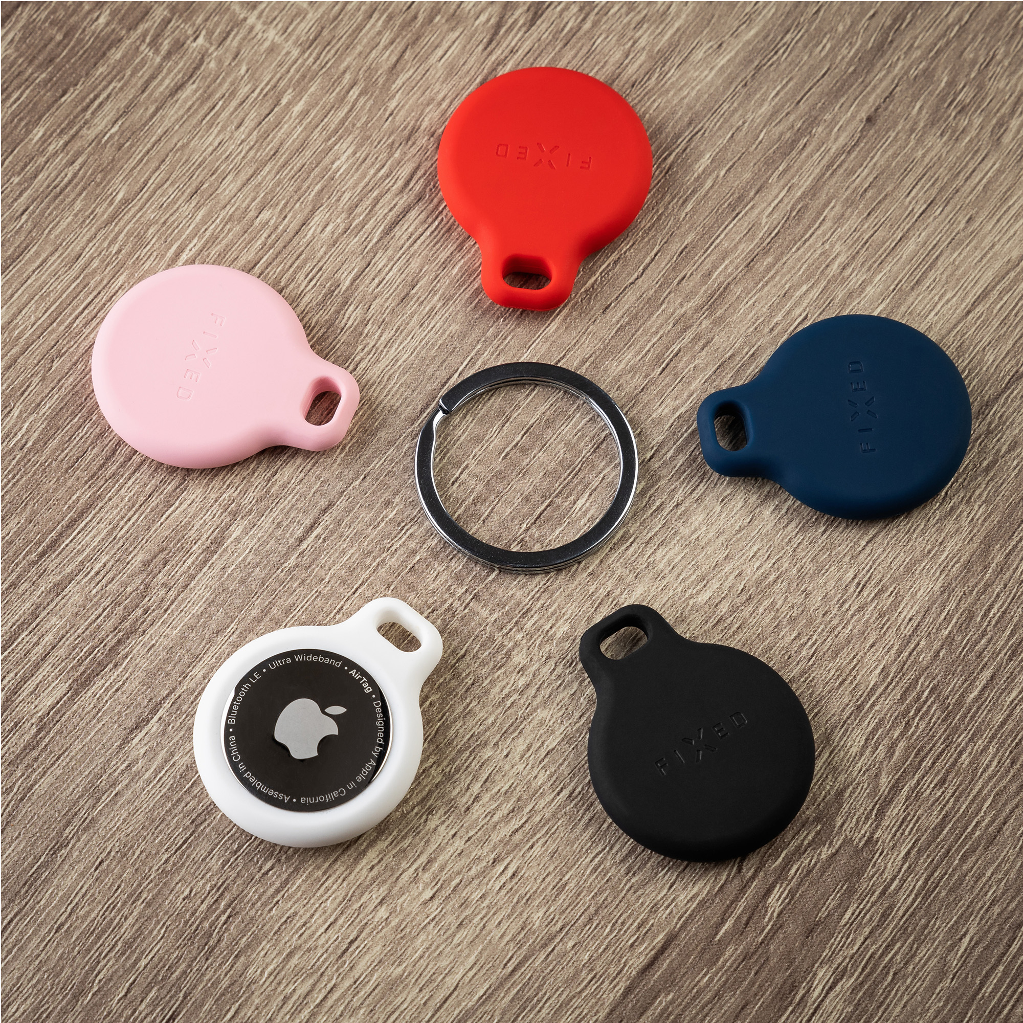 Ein Apple AirTag in weißer Halterung, vier bunte Silikonhüllen und ein Metallschlüsselring auf Holz.