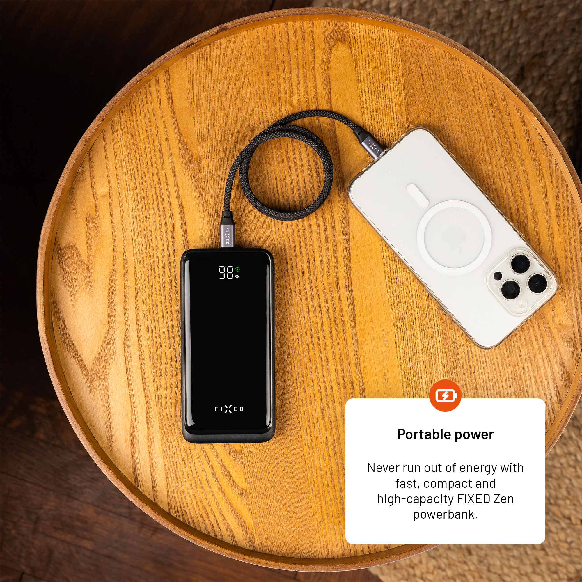 A black powerbank and a white smartphone connected by a cable on a round wooden table.