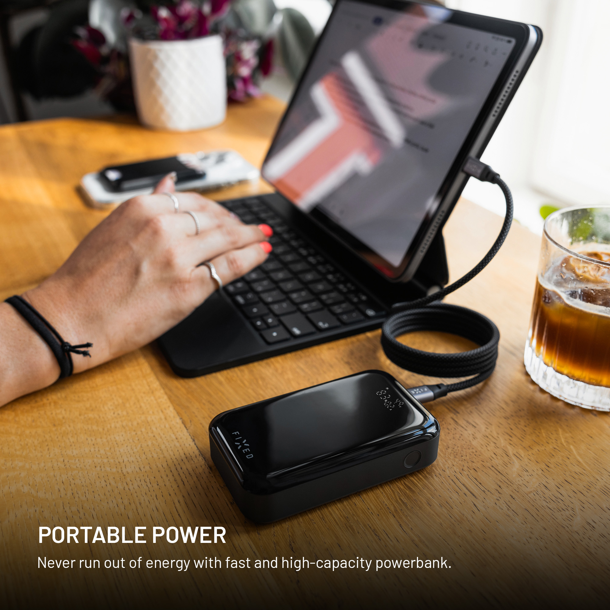 Eine Hand tippt auf einem Tablet, das über ein Kabel mit einer schwarzen Powerbank verbunden ist.