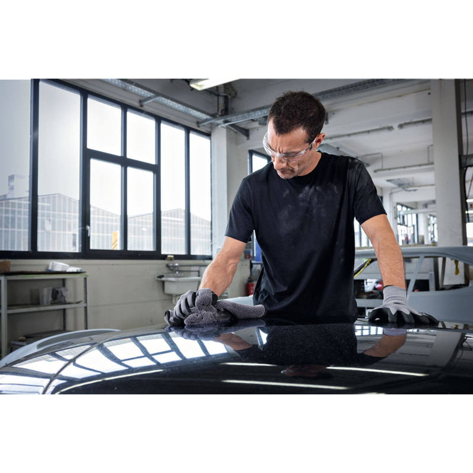 Ein Arbeiter in Schutzbrille und T-Shirt wischt die Motorhaube eines dunklen Autos in einer Werkstatt ab.
